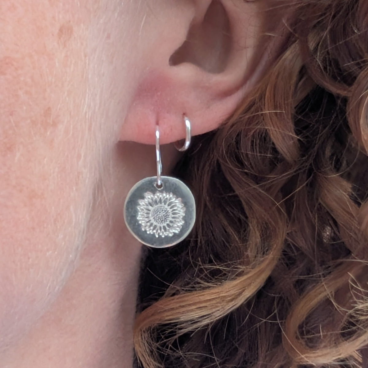 Silver sunflower disc earrings being worn, showing the round sterling silver disc with its detailed stamped sunflower design hanging from a silver hook ear wire.