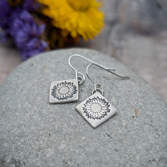 Earring is a diamond shaped plate stamped with a single sunflower, suspended from silver earring wire.