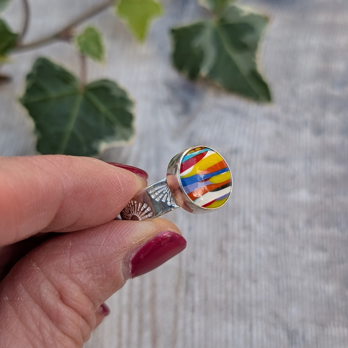 Close up side view of silver sunrise surfite ring held between fingers, highlighting the hand stamped sunrise design on the silver band and colourful Surfite stone.