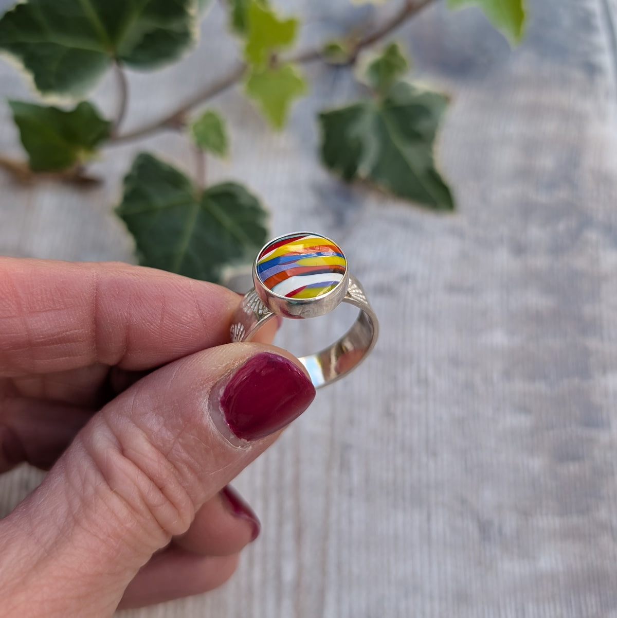 Silver sunrise surfite ring handmade from recycled sterling silver, held between fingers showing the colourful round Surfite stone and hand stamped silver band.