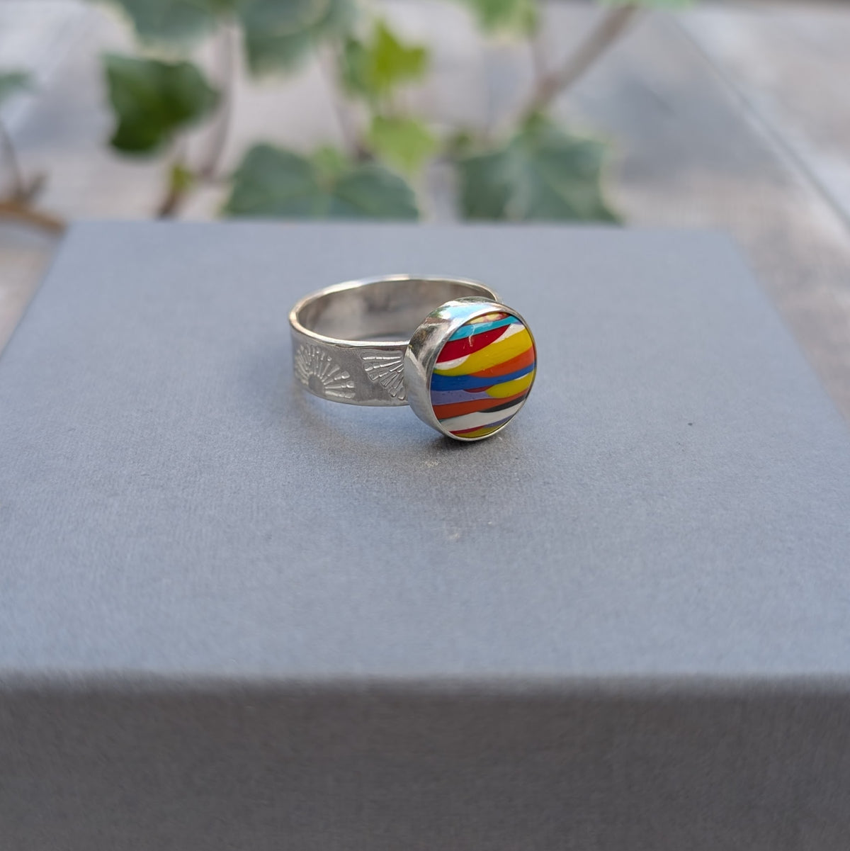 Side view of silver sunrise surfite ring displayed on a grey background, showing the handmade silver bezel setting and hand stamped sunrise pattern on the wide band.