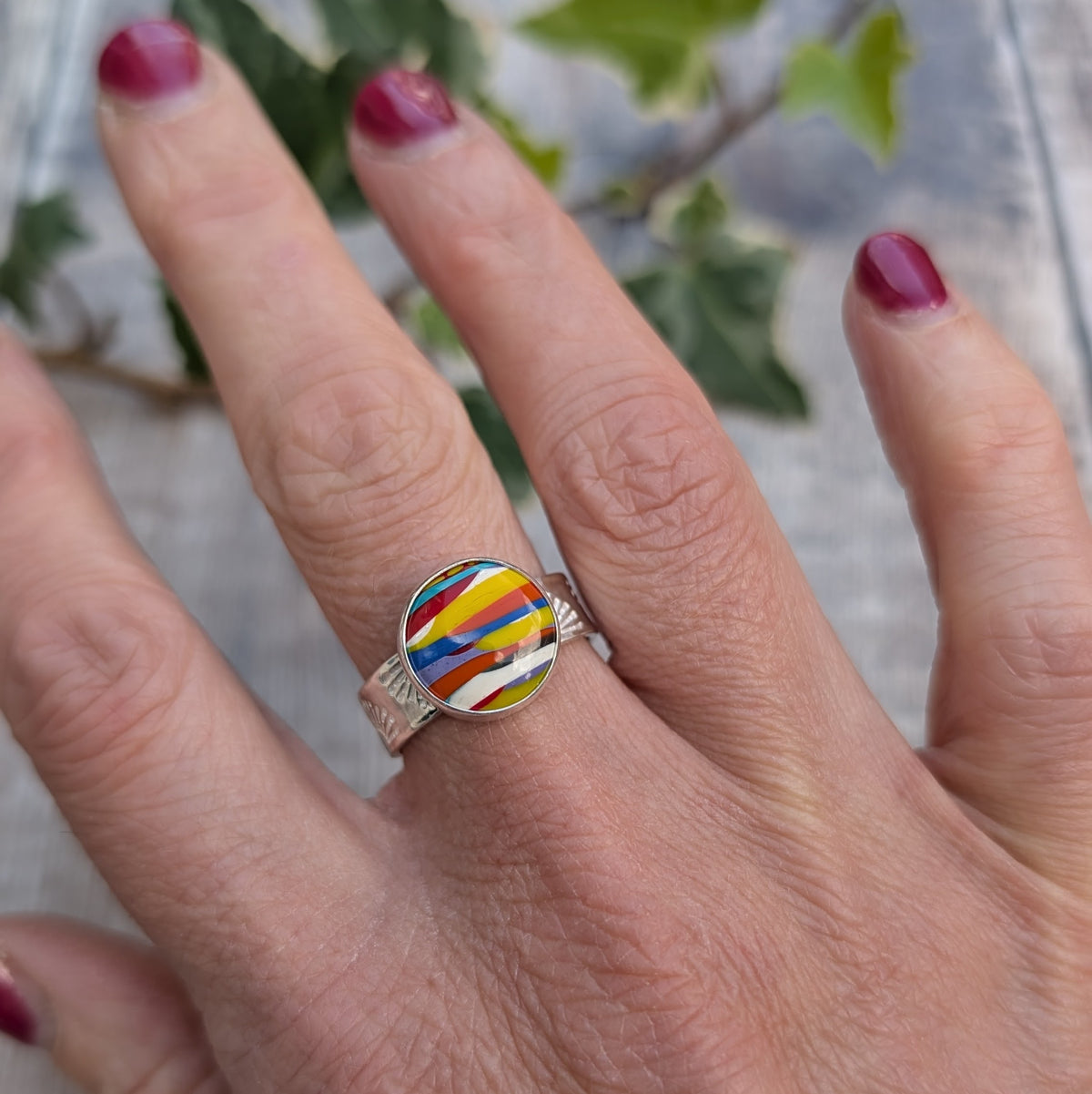 Silver sunrise surfite ring worn on a finger, showing the colourful round Surfite stone set in recycled sterling silver with a patterned band.