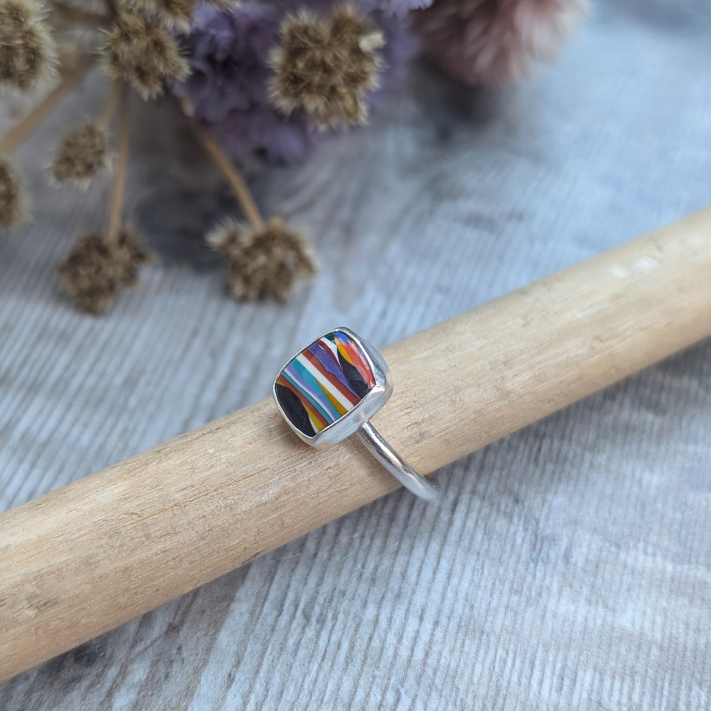 Sterling Silver rectangular shaped surfite ring. Resin stone with rounded edges and gloss surface measuring approximately 10 x 8 mm. Set in a silver bezel and mounted on silver ring band approximately 2 mm wide. Uneven horizonal stripes of colour are mainly orange, blue, black, white, red and yellow.