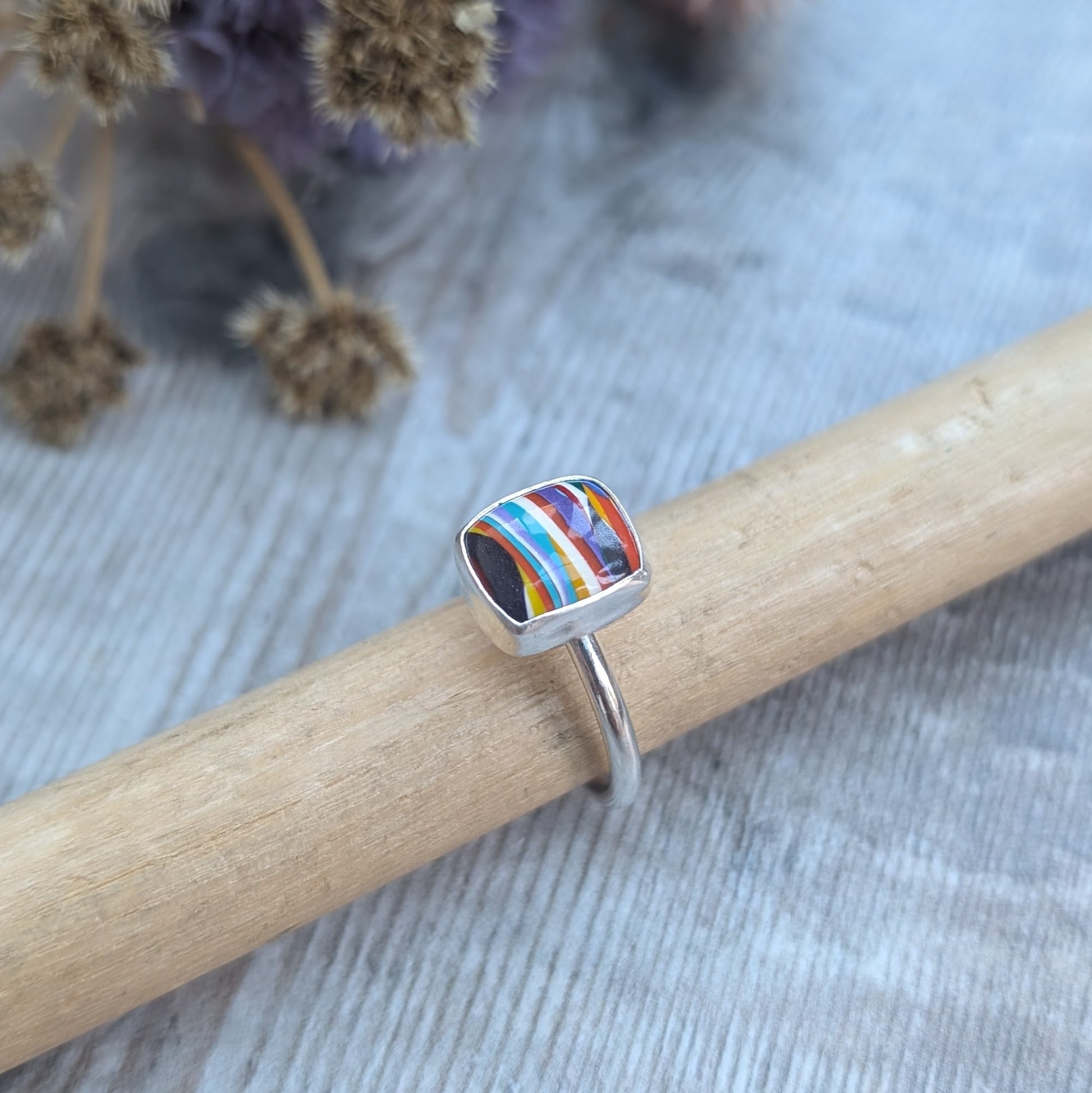 Sterling Silver rectangular shaped surfite ring. Resin stone with rounded edges and gloss surface measuring approximately 10 x 8 mm. Set in a silver bezel and mounted on silver ring band approximately 2 mm wide. Uneven horizonal stripes of colour are mainly orange, blue, black, white, red and yellow.