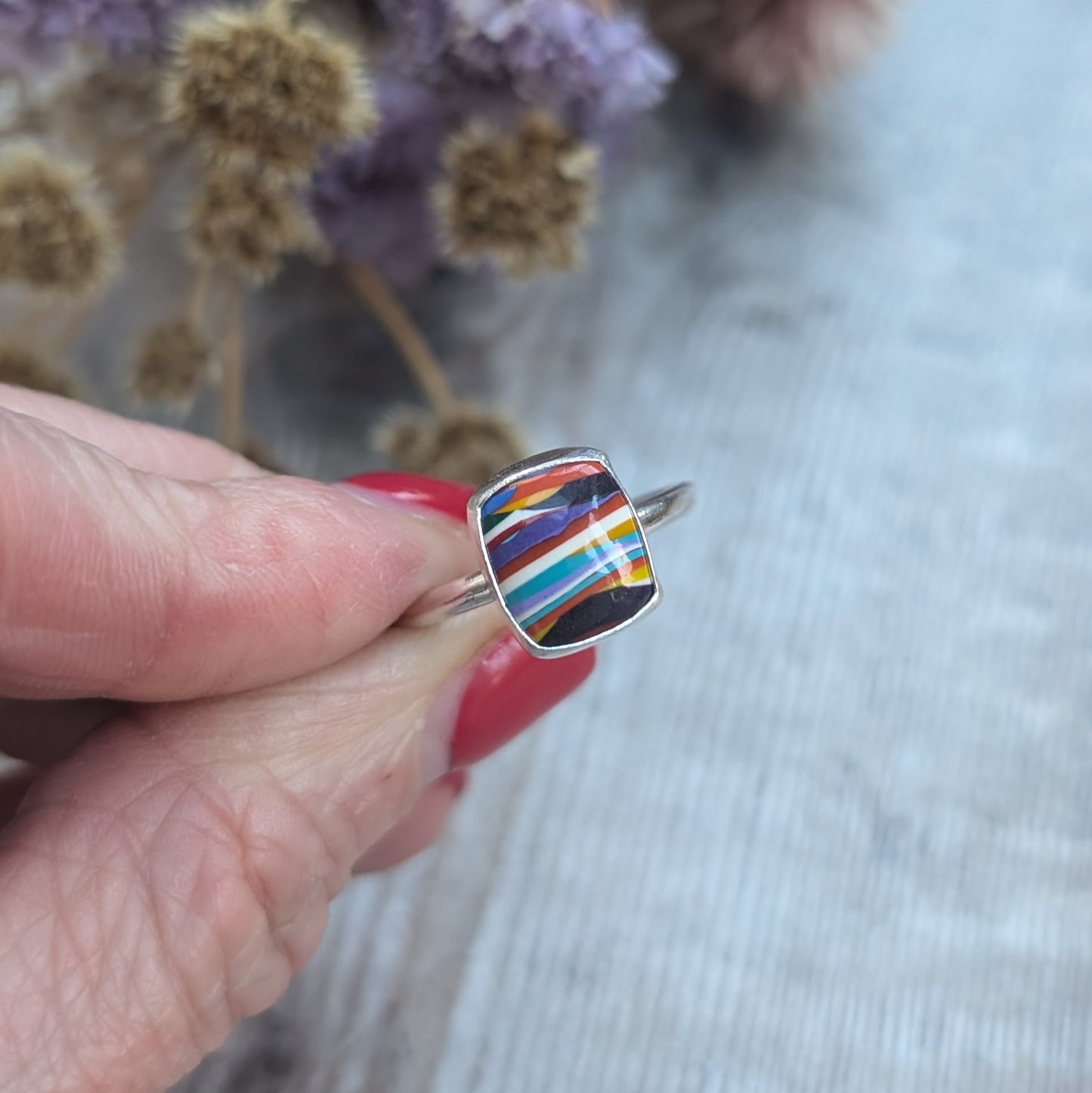 Sterling Silver rectangular shaped surfite ring. Resin stone with rounded edges and gloss surface measuring approximately 10 x 8 mm. Set in a silver bezel and mounted on silver ring band approximately 2 mm wide. Uneven horizonal stripes of colour are mainly orange, blue, black, white, red and yellow.