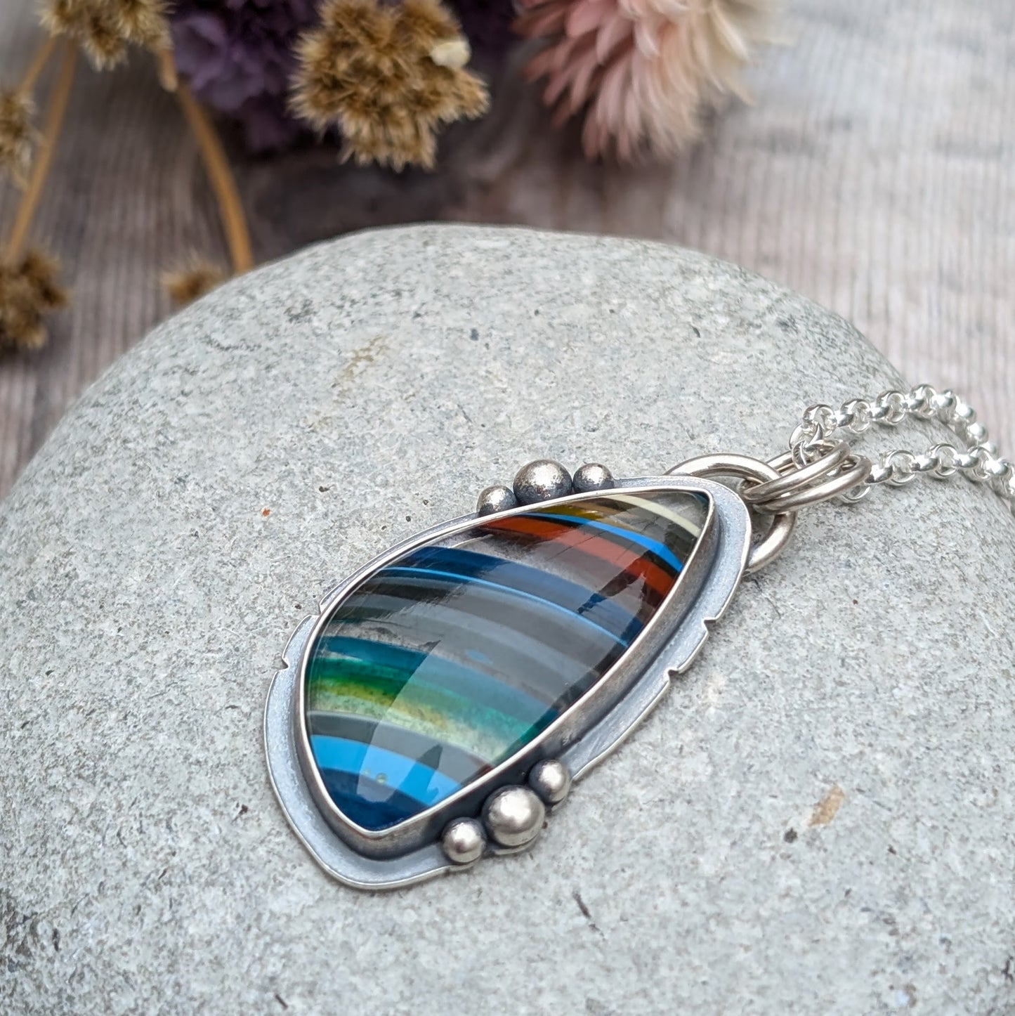 Close-up of a Surfite necklace with a colourful striped Surfite stone, framed in oxidised sterling silver with decorative pebble details, resting on a smooth stone.