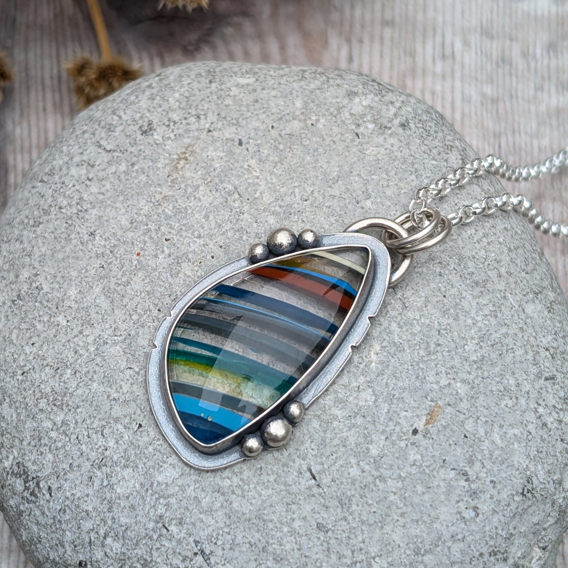 Close-up of a Surfite necklace with a colourful striped Surfite stone, framed in oxidised sterling silver with decorative pebble details, resting on a smooth stone.