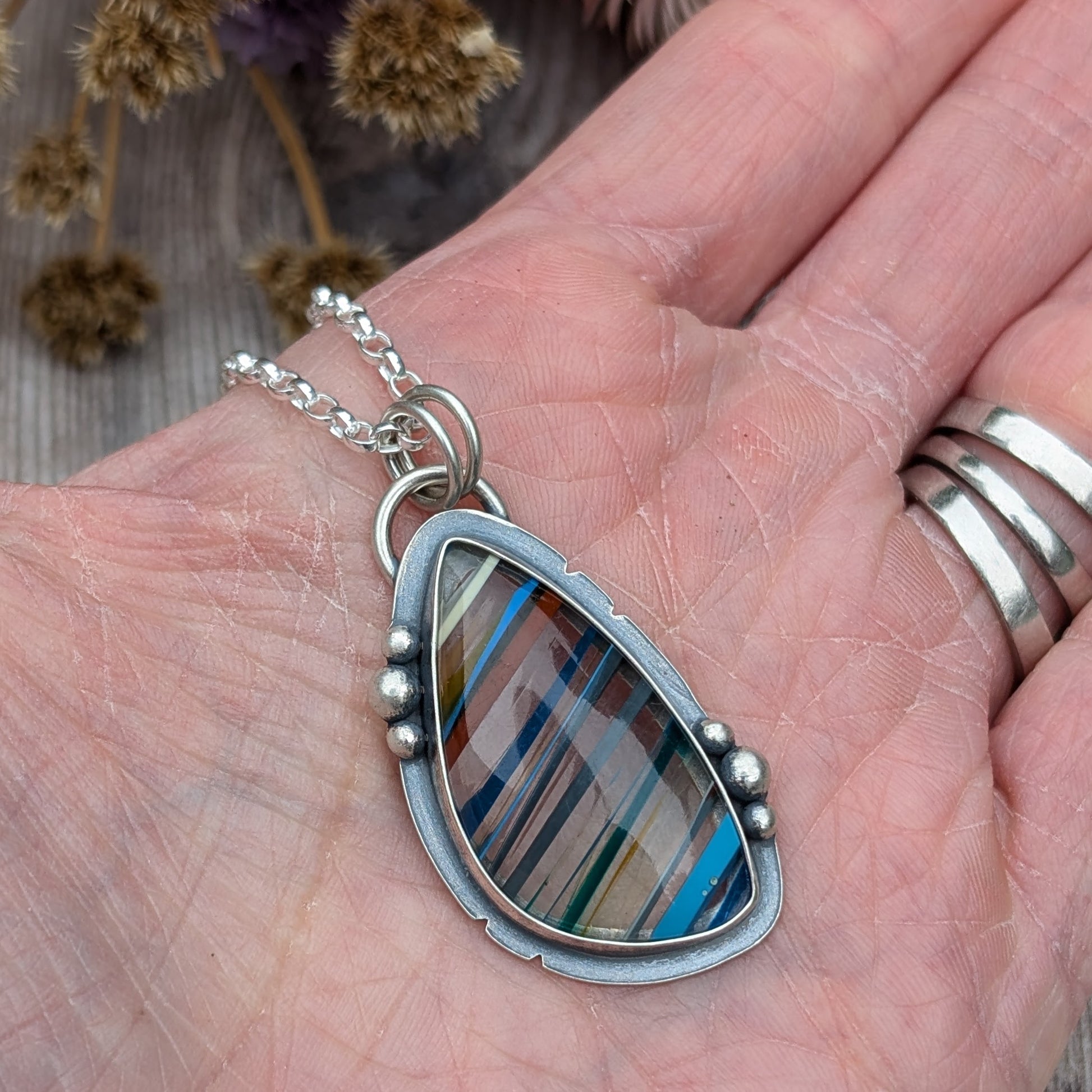 Handheld Surfite necklace showing the vibrant, multi-coloured Surfite stone set in oxidised silver, accented by small pebble details on the sides of the pendant.