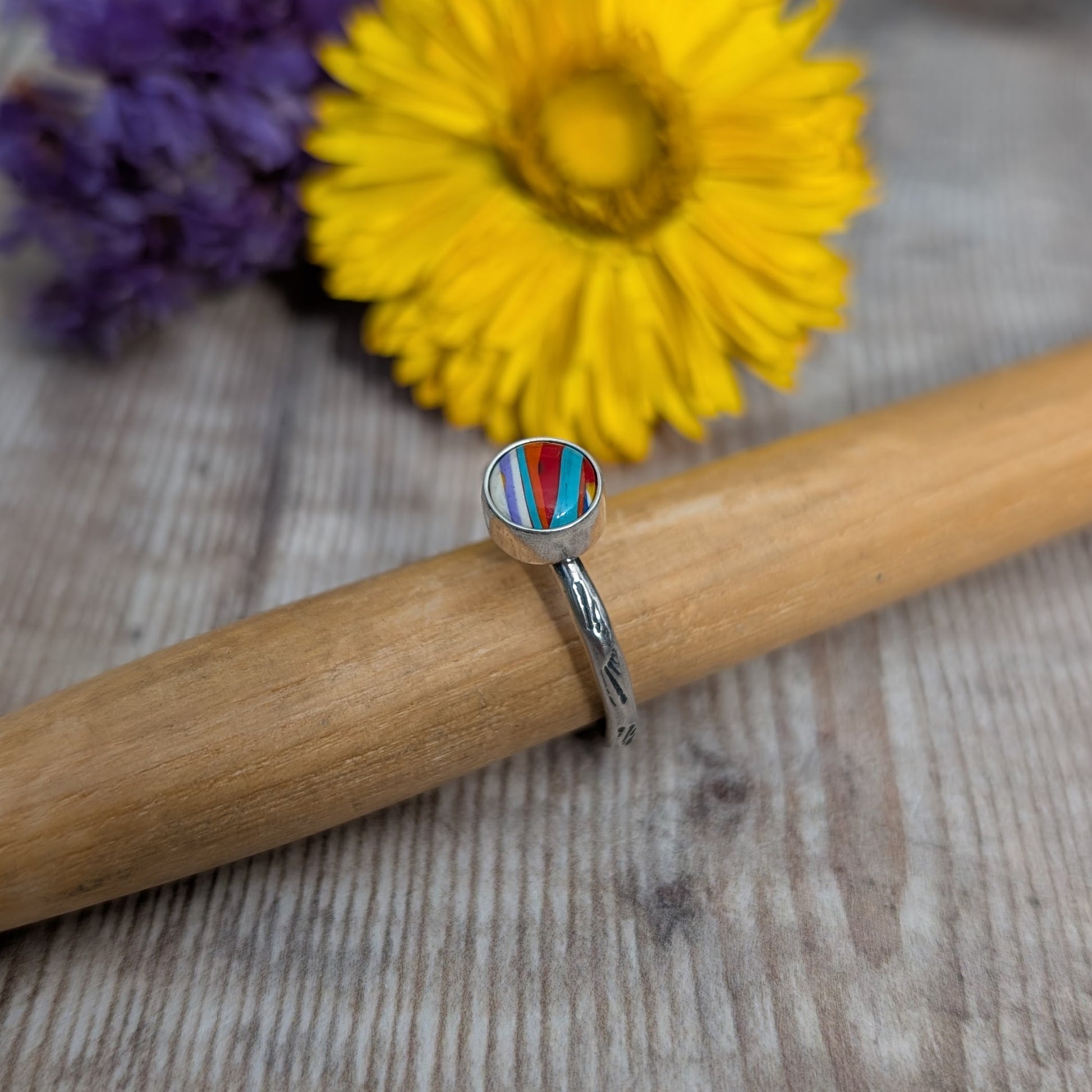 Surfite Ring with Sunburst Stamp close-up on a mandrel with flowers in the background, showing the handmade setting and striped Surfite stone.