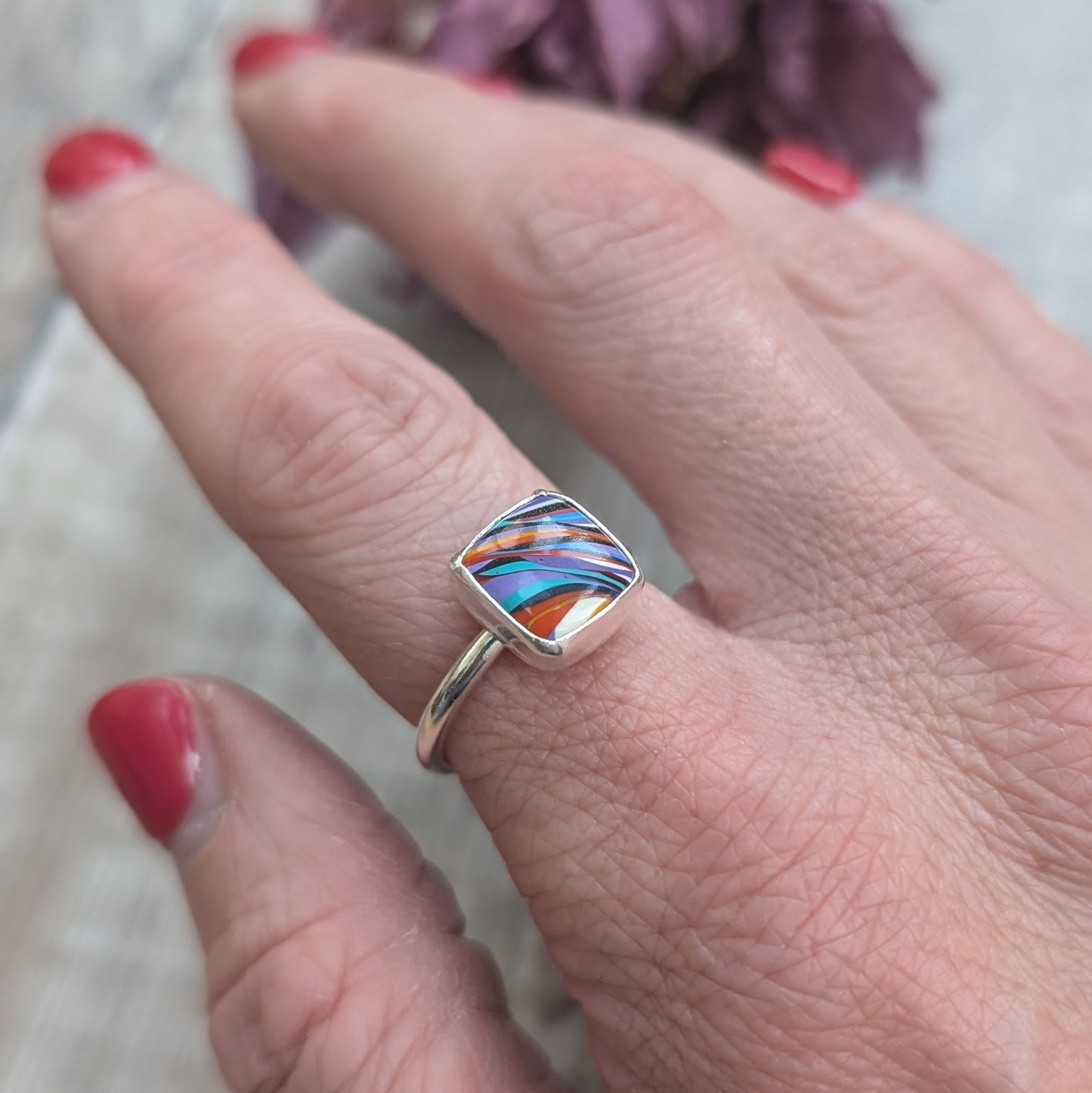 Sterling Silver square surfite resin stone with rounded edges and gloss surface measuring approximately 9 x 9mm. Set in a silver bezel and mounted on silver ring band approx 2 mm wide. Uneven diagonal stripes of colour are mainly orange, blue, black, white and yellow.