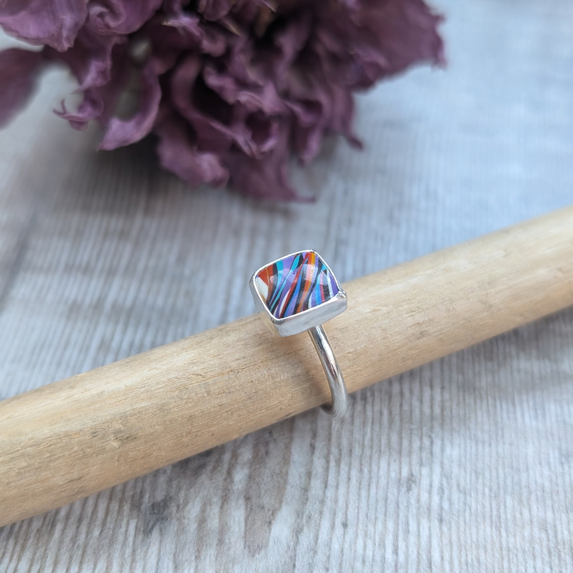 Sterling Silver square surfite resin stone with rounded edges and gloss surface measuring approximately 9 x 9mm. Set in a silver bezel and mounted on silver ring band approx 2 mm wide. Uneven diagonal stripes of colour are mainly orange, blue, black, white and yellow.