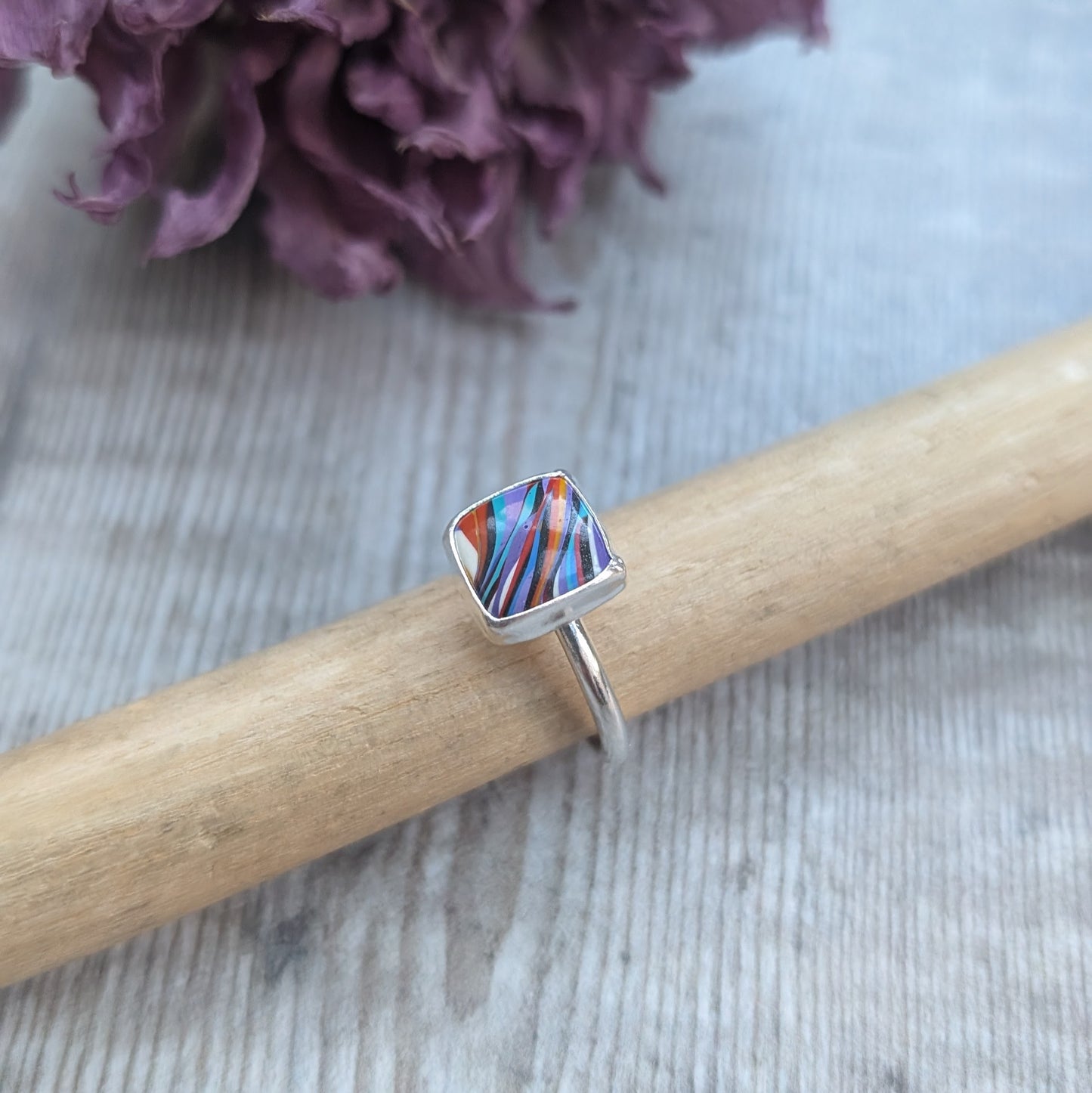 Sterling Silver square surfite resin stone with rounded edges and gloss surface measuring approximately 9 x 9mm. Set in a silver bezel and mounted on silver ring band approx 2 mm wide. Uneven diagonal stripes of colour are mainly orange, blue, black, white and yellow.