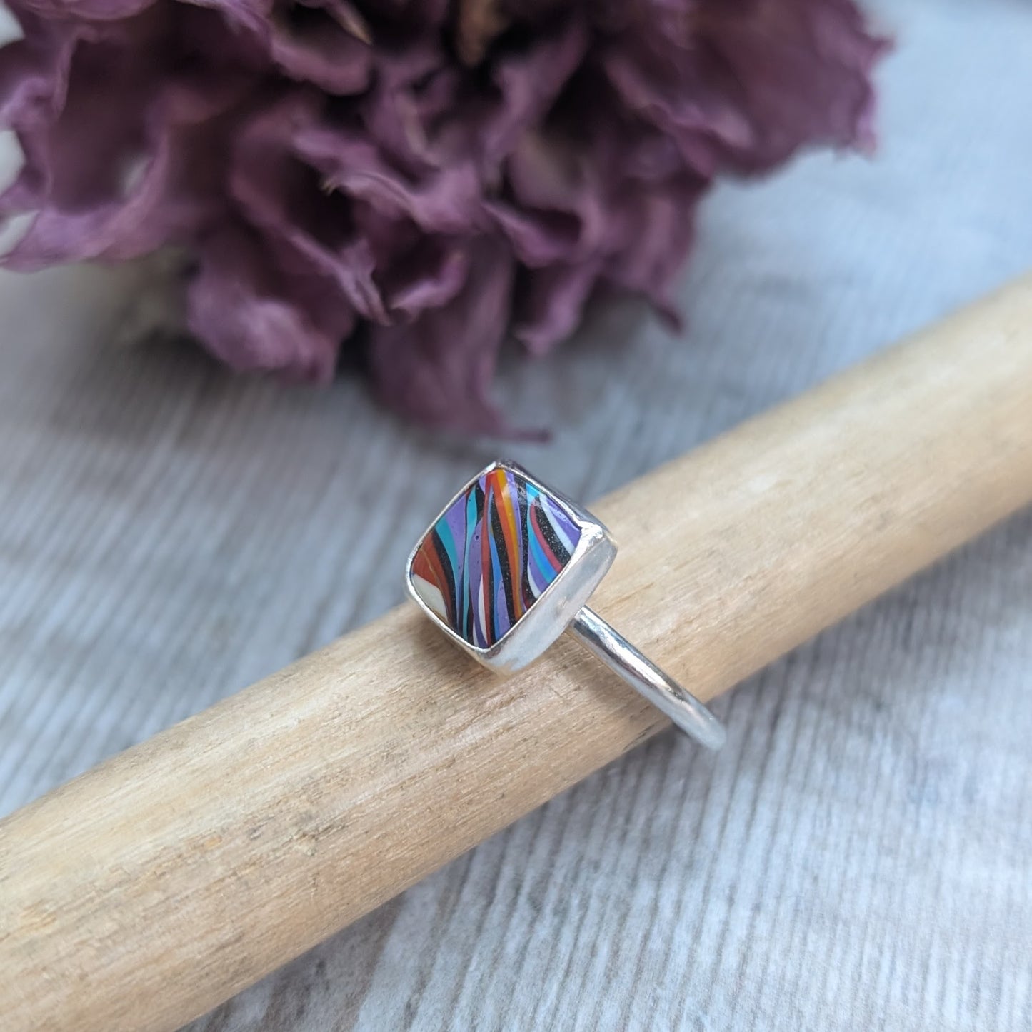 Sterling Silver square surfite resin stone with rounded edges and gloss surface measuring approximately 9 x 9mm. Set in a silver bezel and mounted on silver ring band approx 2 mm wide. Uneven diagonal stripes of colour are mainly orange, blue, black, white and yellow.