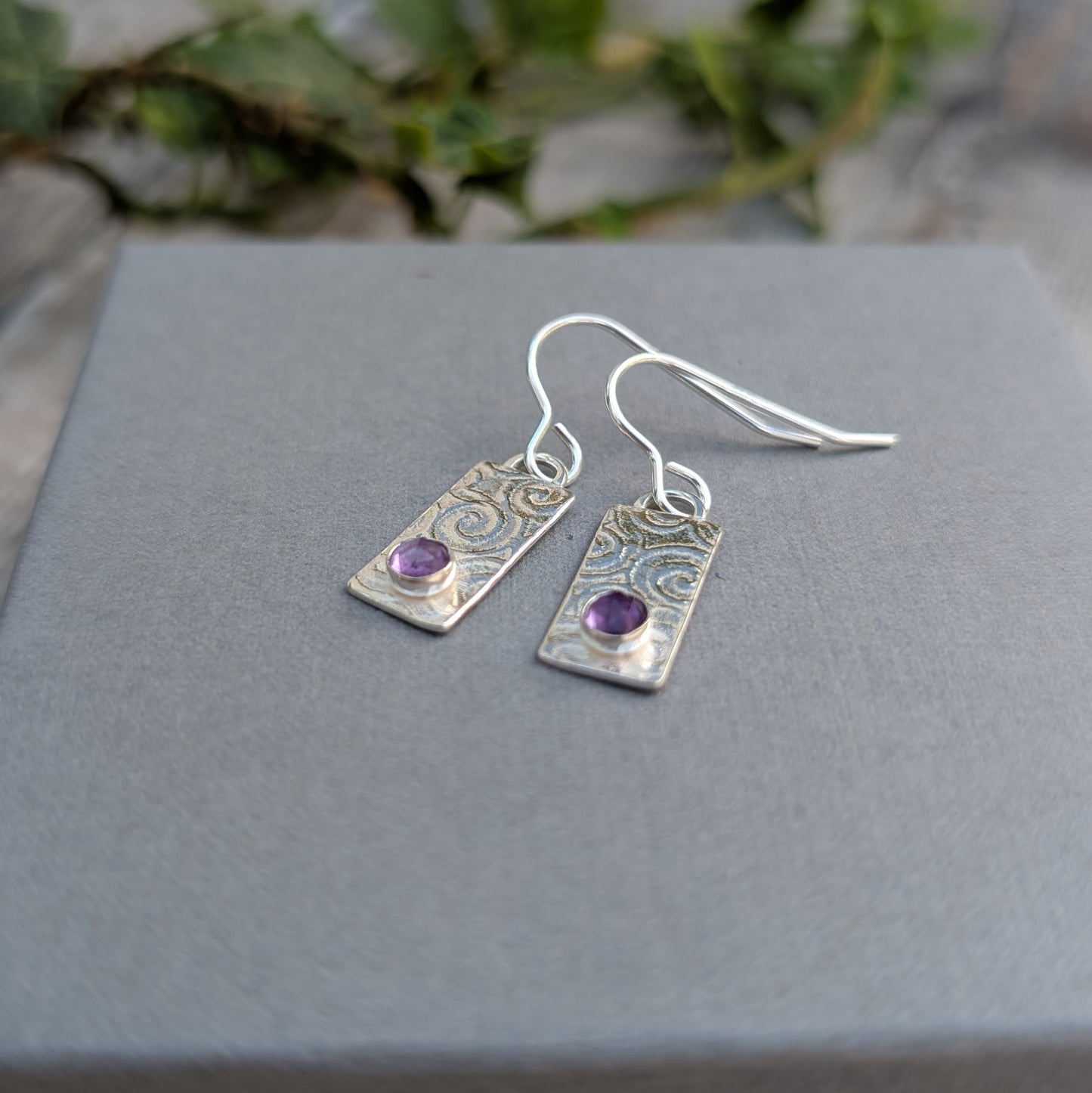 Swirly Rectangle Patterned Earrings with Amethyst made from recycled Sterling Silver, photographed on a grey display card, showing the hand-stamped swirly pattern and faceted Amethyst stones.