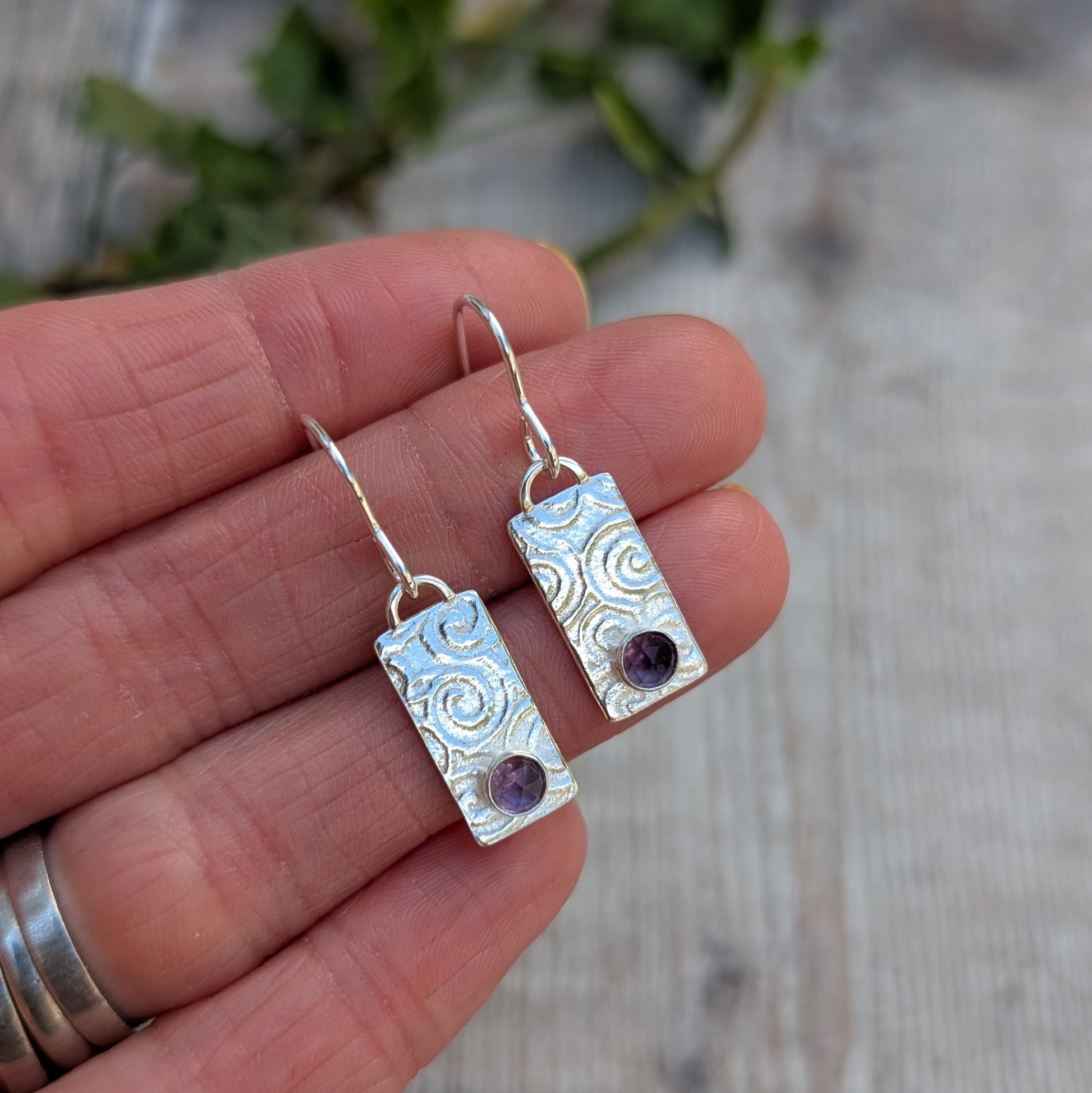 Swirly Rectangle Patterned Earrings with Amethyst handmade from recycled Sterling Silver, held in a hand to show size, featuring a hand-stamped swirly pattern and round purple gemstones.