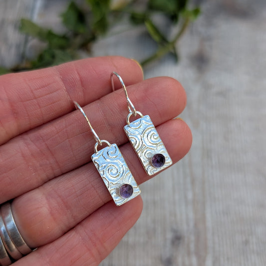 Swirly Rectangle Patterned Earrings with Amethyst handmade from recycled Sterling Silver, held in a hand to show size, featuring a hand-stamped swirly pattern and round purple gemstones.