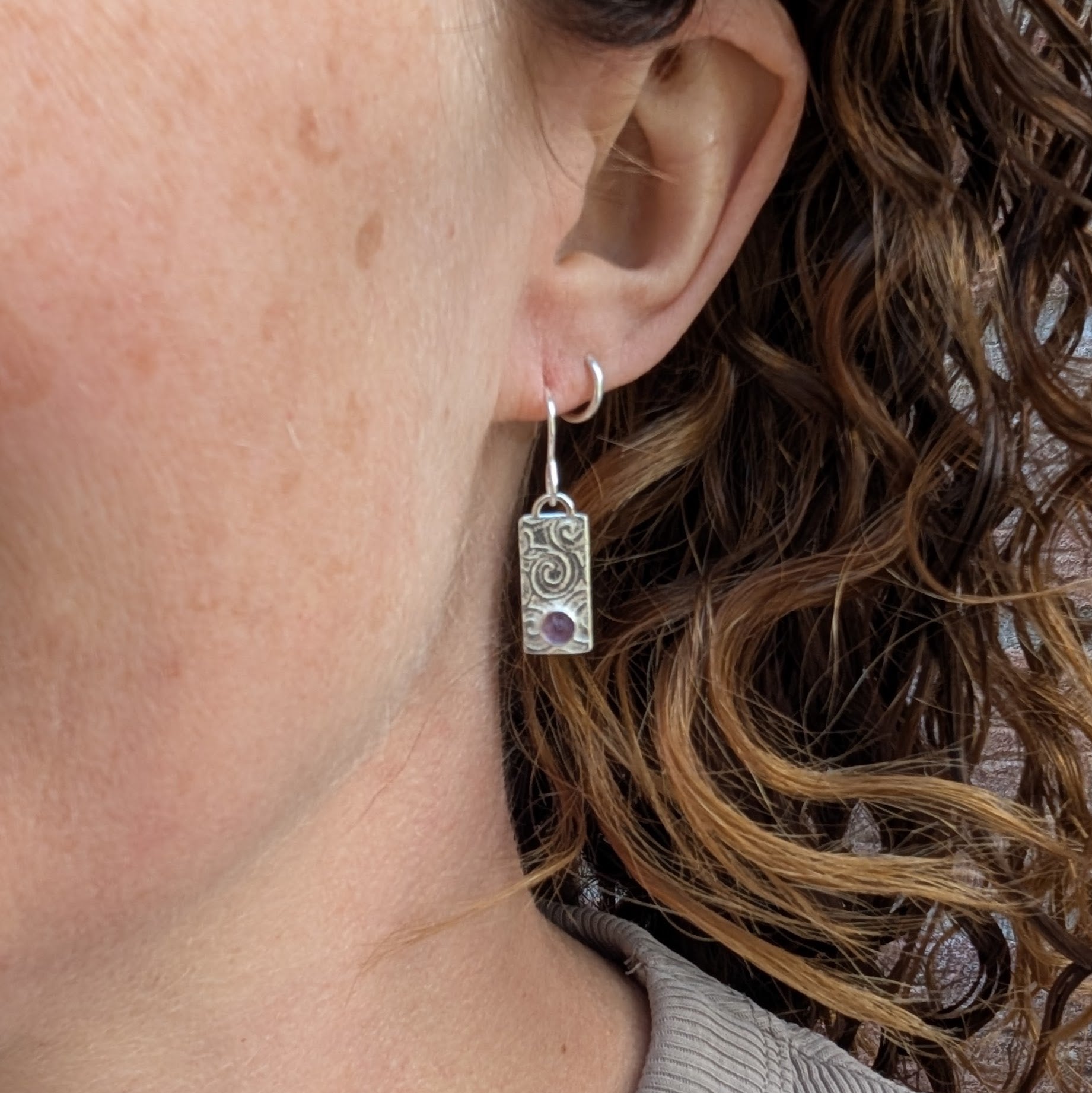 Swirly Rectangle Patterned Earrings with Amethyst worn in an ear, showing the rectangular recycled Sterling Silver drops, hand-stamped swirly pattern and purple Amethyst stones.