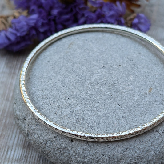 A handmade sterling silver bangle with a hammered texture is displayed resting on a smooth grey stone. The subtle light reflections highlight the intricate hammered details of the bangle, with soft purple and yellow flowers in the background.