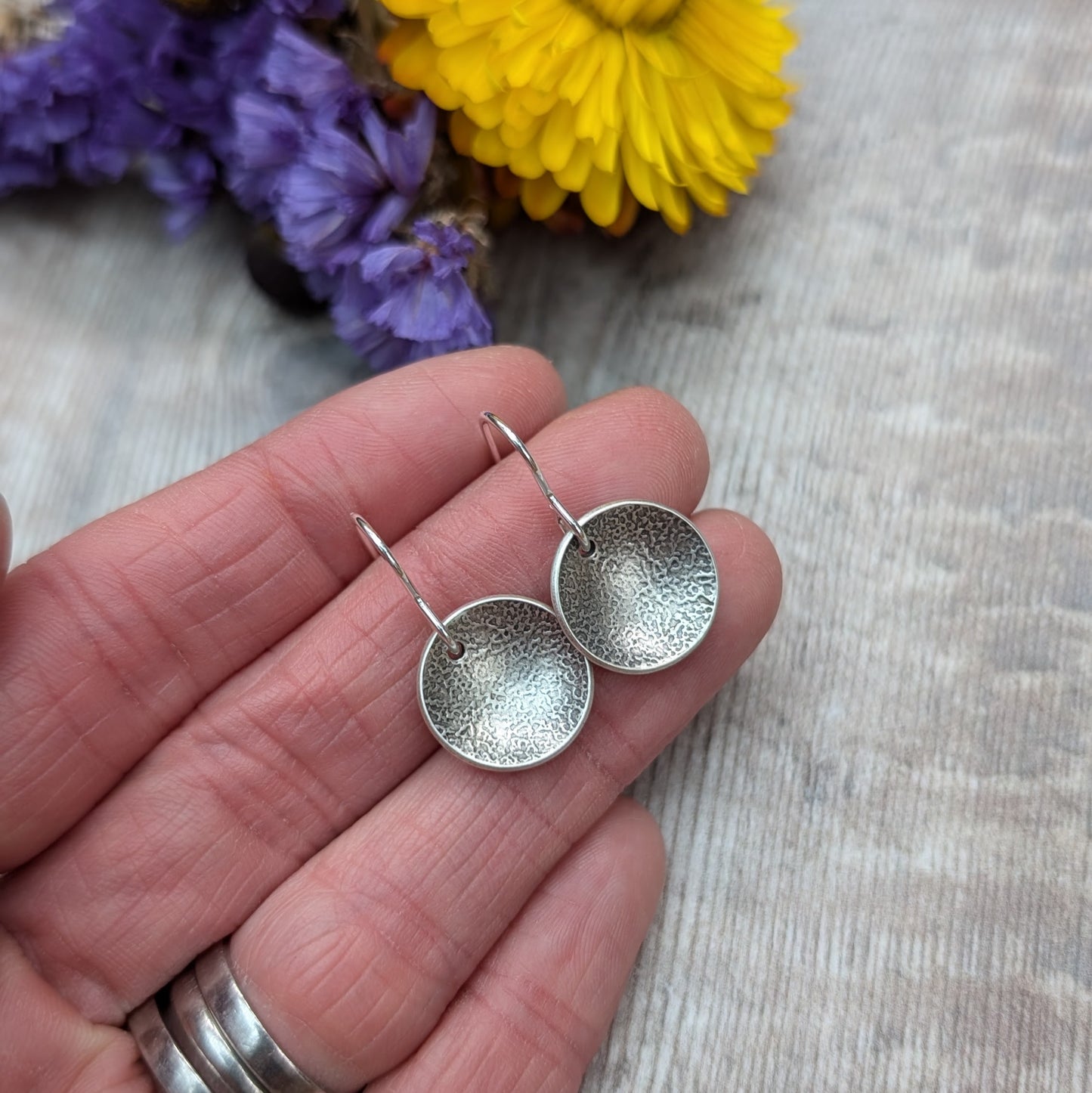 Hand holding a pair of oxidised, recycled sterling silver textured disc earrings with purple and yellow flowers in the background.