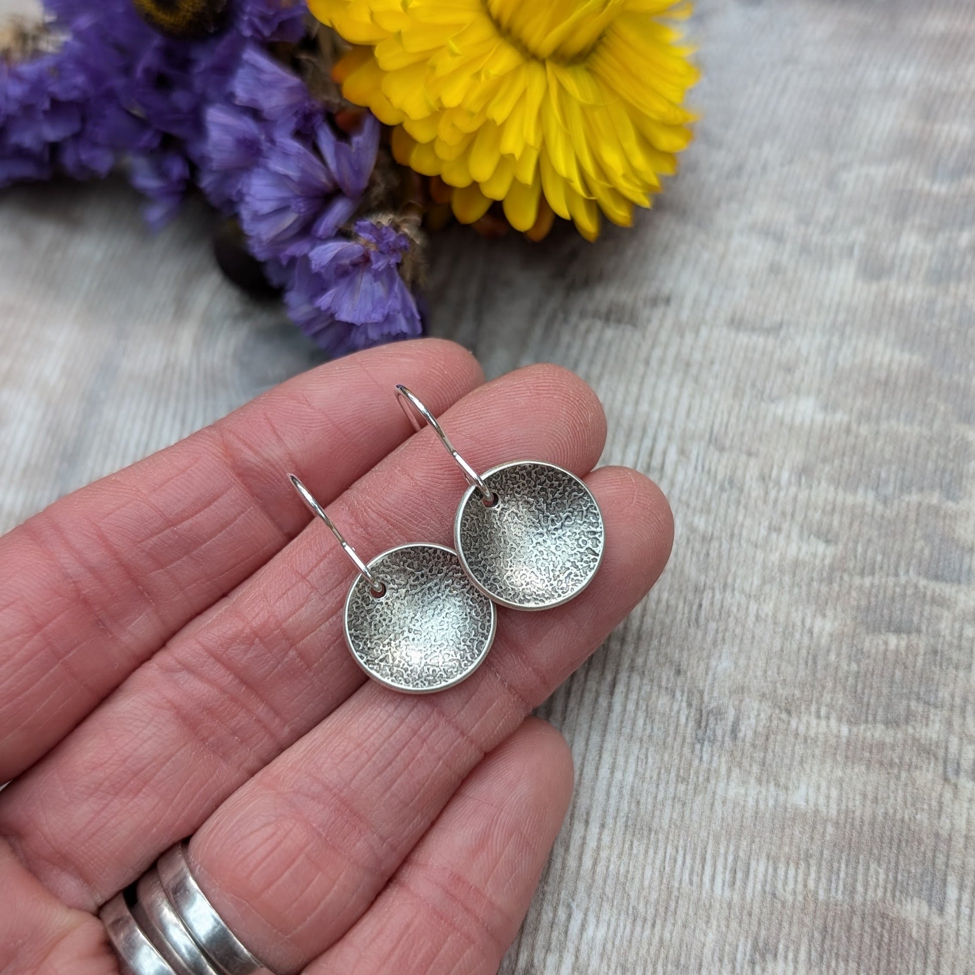 Hand holding a pair of oxidised, recycled sterling silver textured disc earrings with purple and yellow flowers in the background.