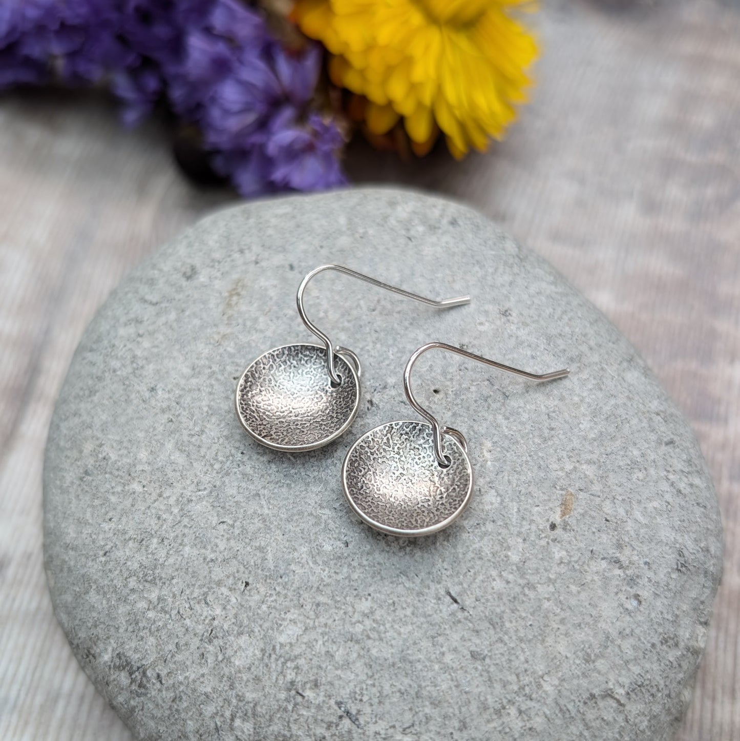 Close-up of oxidised, recycled sterling silver textured disc earrings on a gray stone with flowers in the background.