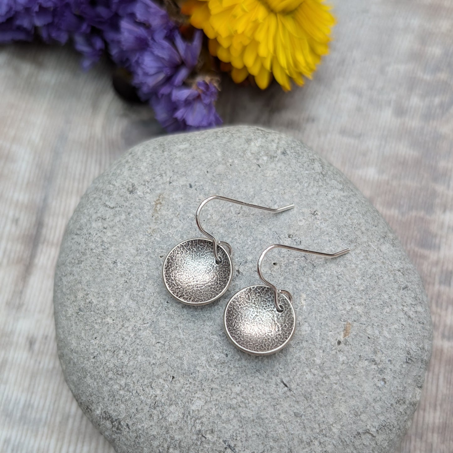 Close-up of oxidised, recycled sterling silver textured disc earrings on a gray stone with flowers in the background.