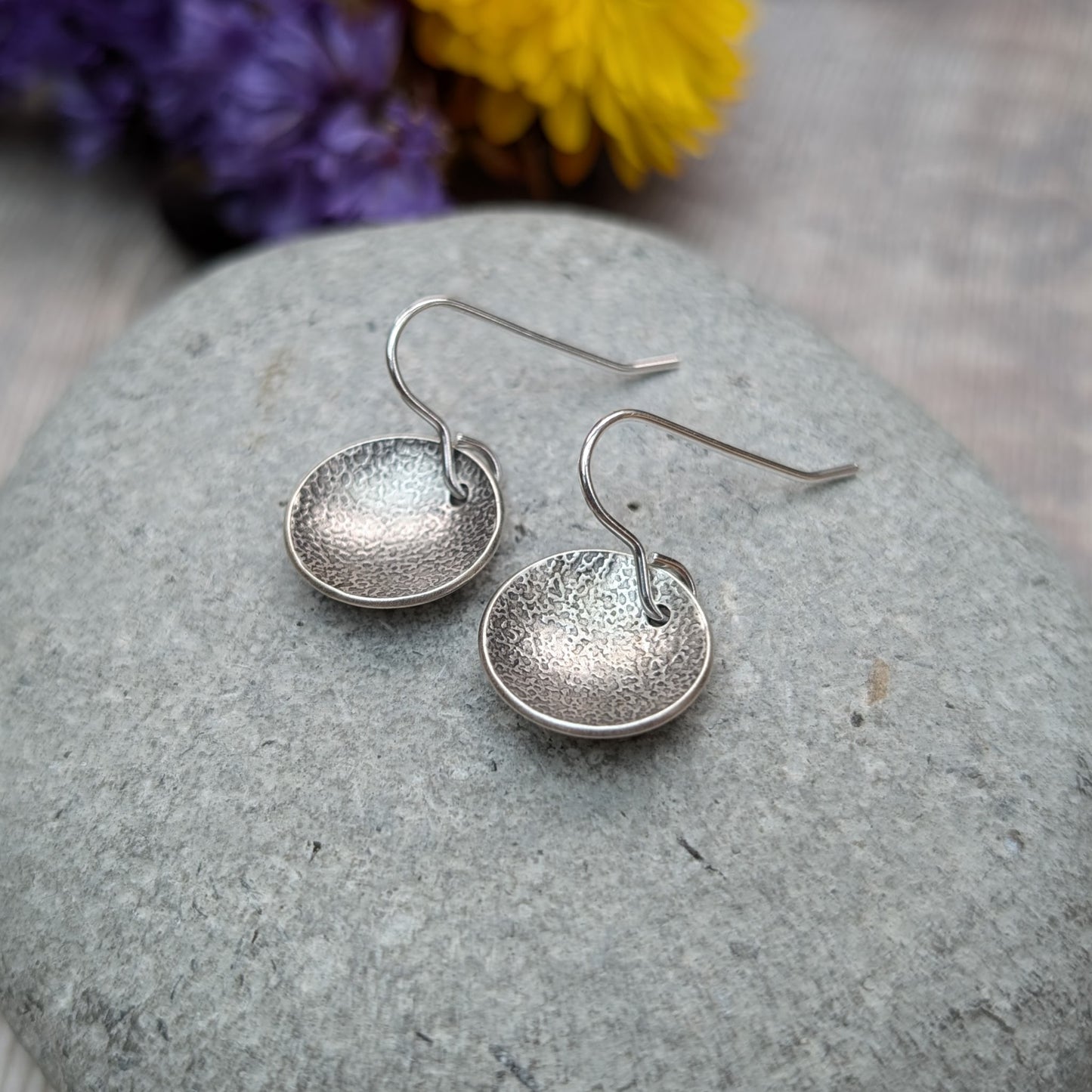 Close-up of oxidised, recycled sterling silver textured disc earrings on a gray stone with flowers in the background.