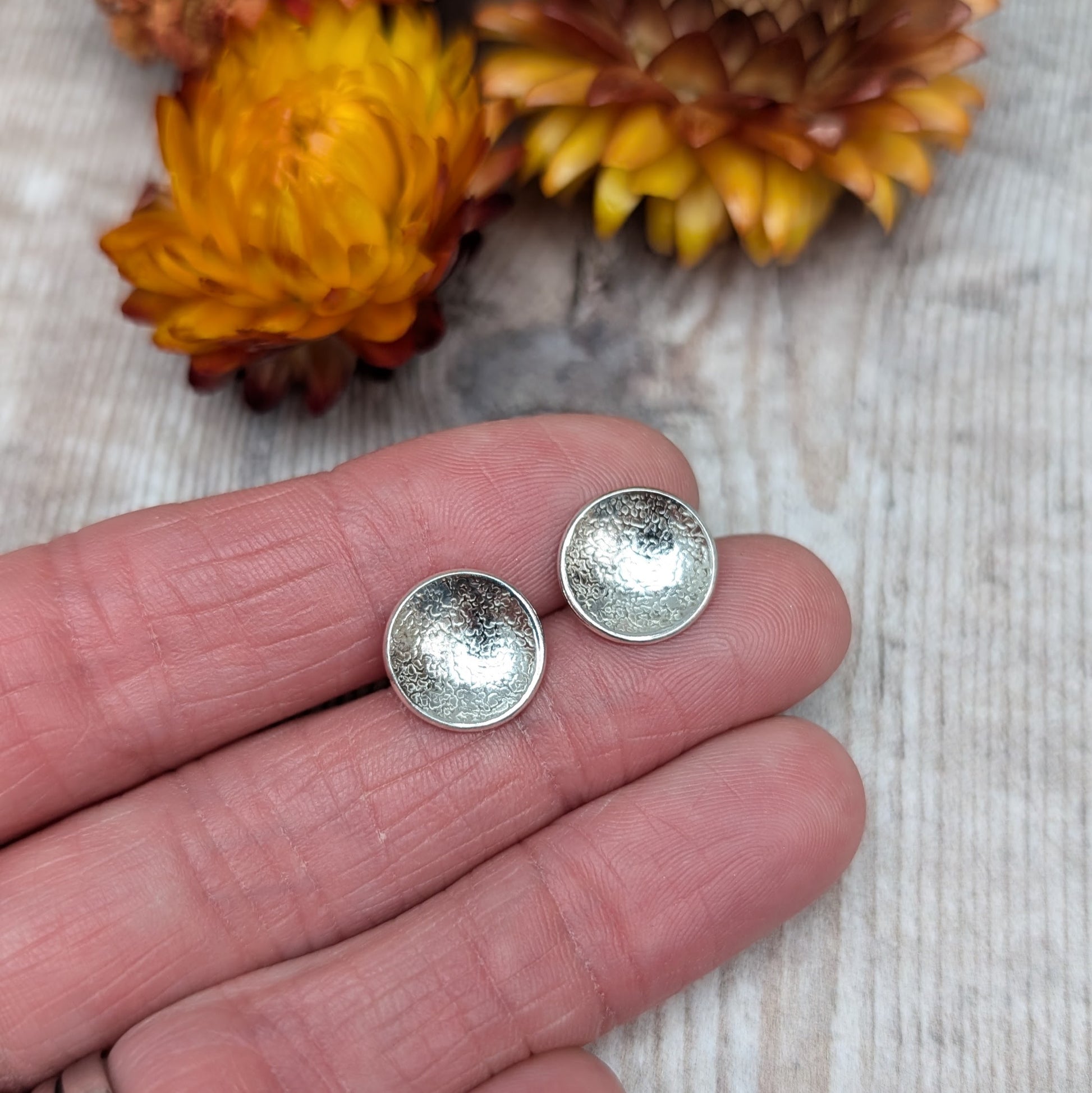 Close-up view of textured domed disc stud earrings in recycled Sterling Silver, held in a hand against a light wooden background with yellow and purple flowers in the background.