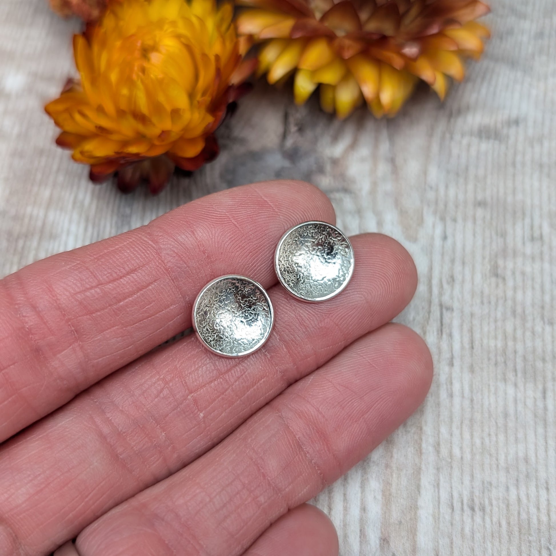 Close-up view of textured domed disc stud earrings in recycled Sterling Silver, held in a hand against a light wooden background with yellow and purple flowers in the background.