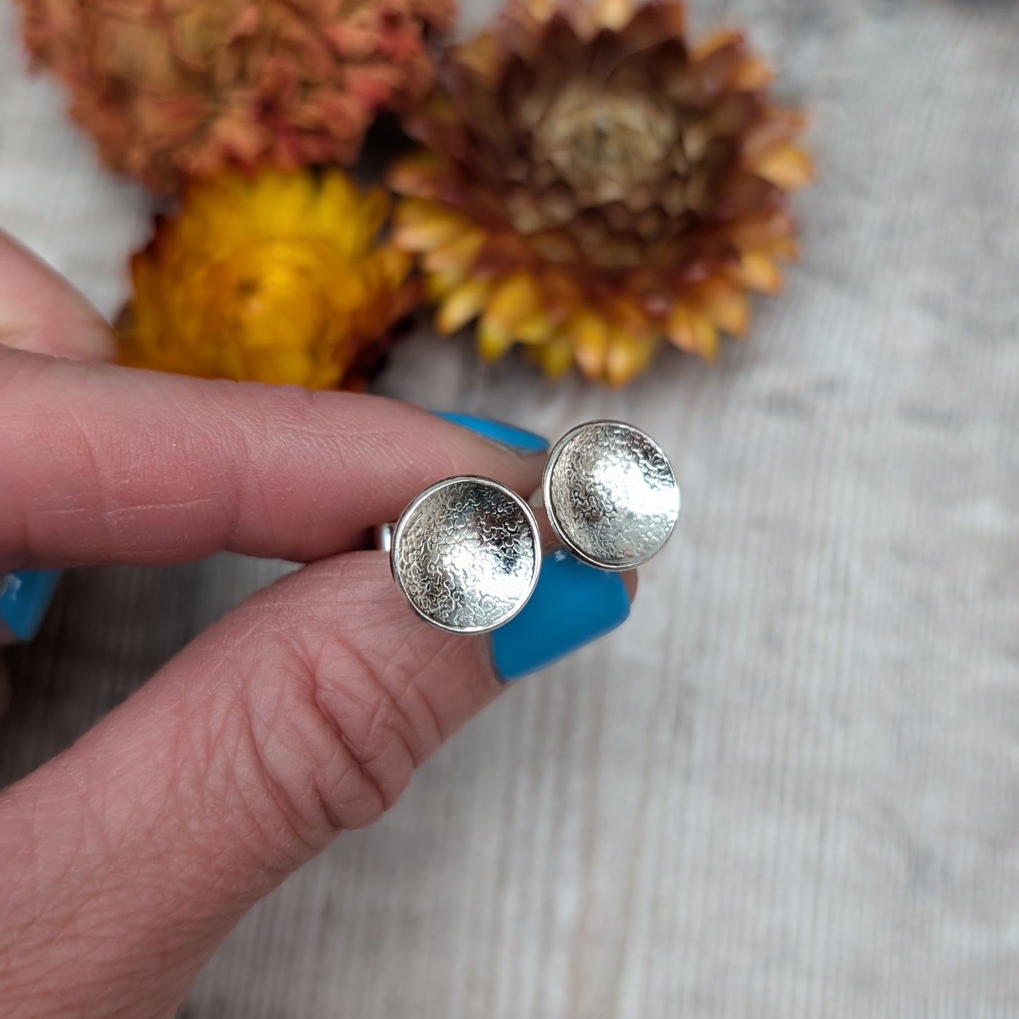 Close-up view of textured domed disc stud earrings in recycled Sterling Silver, held in a hand against a light wooden background with yellow and purple flowers in the background.