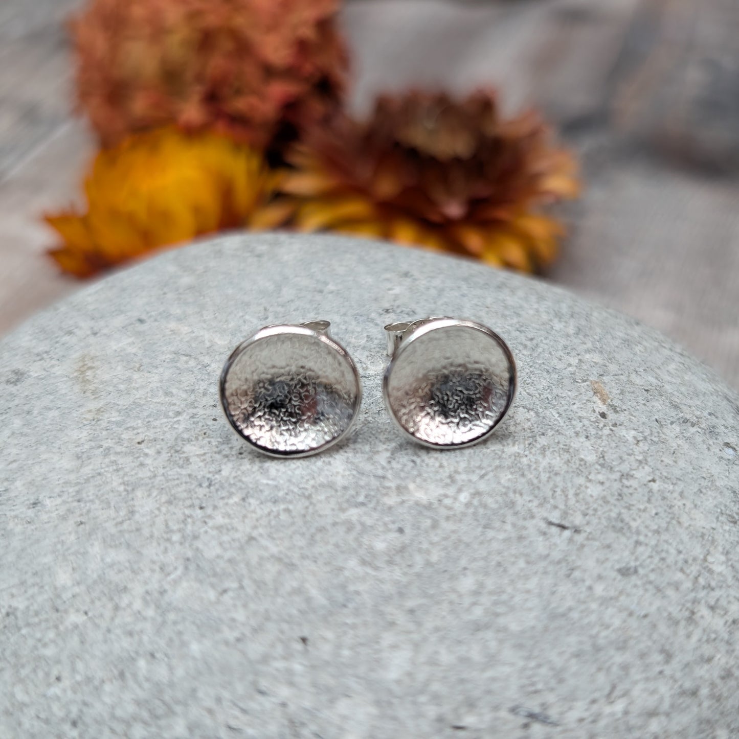 Pair of textured domed disc stud earrings made from recycled Sterling Silver, displayed on a smooth stone with blurred yellow and brown flowers in the background.