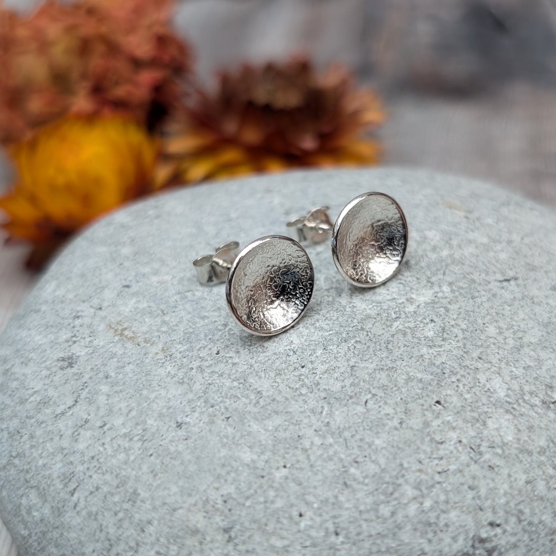 Pair of textured domed disc stud earrings made from recycled Sterling Silver, displayed on a smooth stone with blurred yellow and brown flowers in the background.