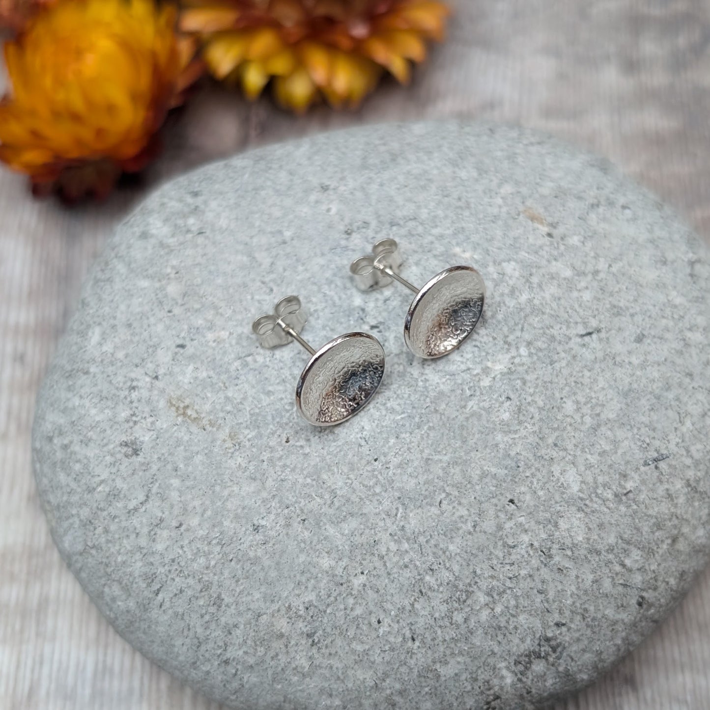 Pair of textured domed disc stud earrings made from recycled Sterling Silver, displayed on a smooth stone with blurred yellow and brown flowers in the background.