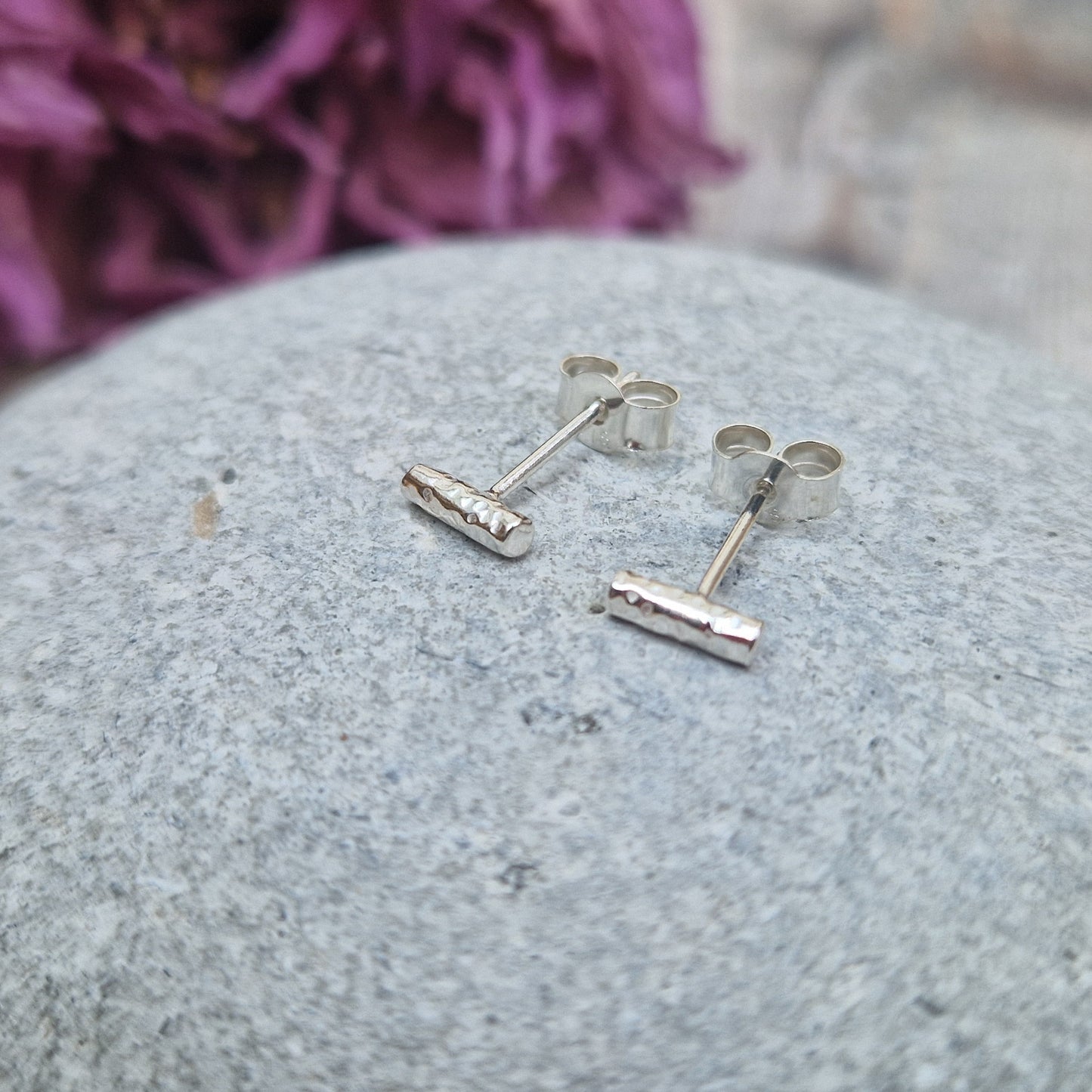 Pair of tiny sterling silver stud earrings with a hammered twig design, displayed on a smooth grey stone with a soft floral background.