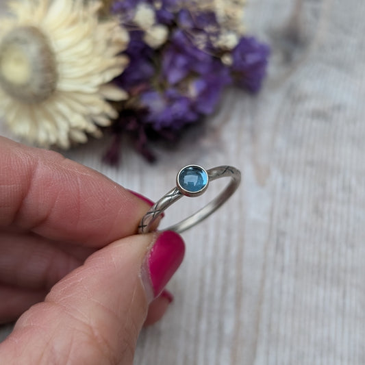 Slim silver band with cross stamped pattern. A blue topaz round stone is mounted on the band in an oxidised sterling silver bezel. Rings can be stacked above or below this piece.