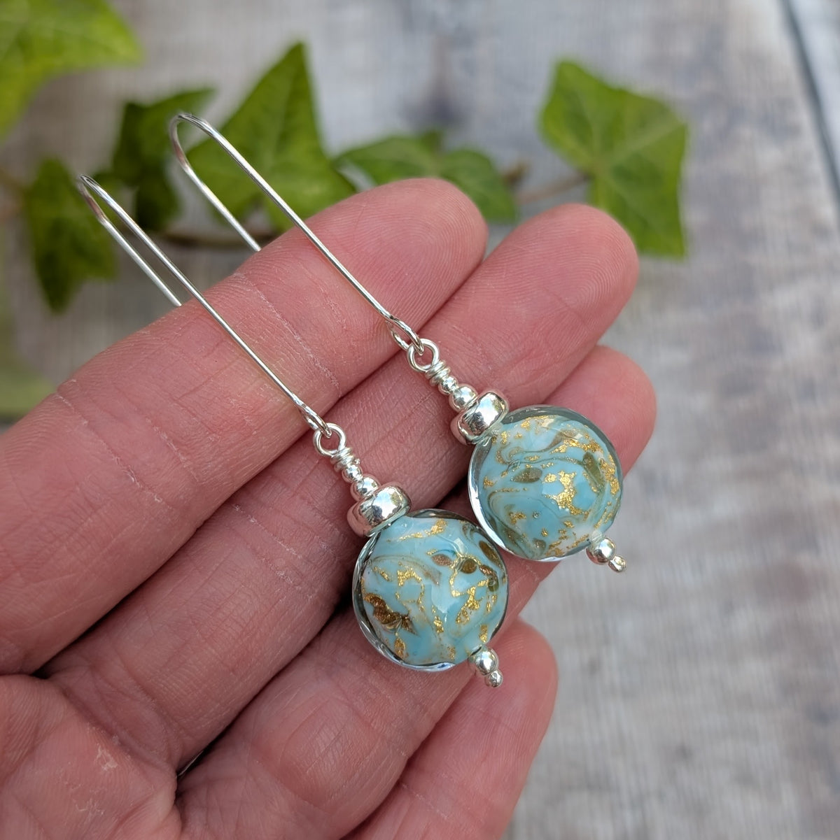 Close up of turquoise marbled Venetian glass long silver earrings held in a hand, with turquoise blue and gold handmade lampwork glass beads.