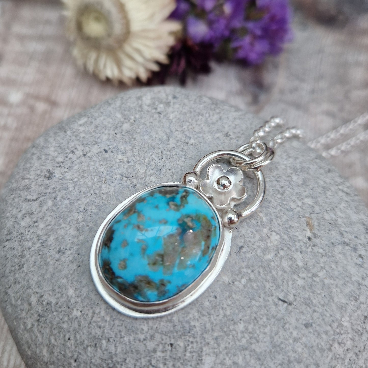 Turquoise oval gemstone pendant set in simple Sterling Silver bezel surround. Fused to the top of the bezel setting is a silver half circle with a five petaled flower in the middle, attached to silver chain via three small silver circles. Gemstone is predominantly turquoise blue with dark, almost black plume formations.