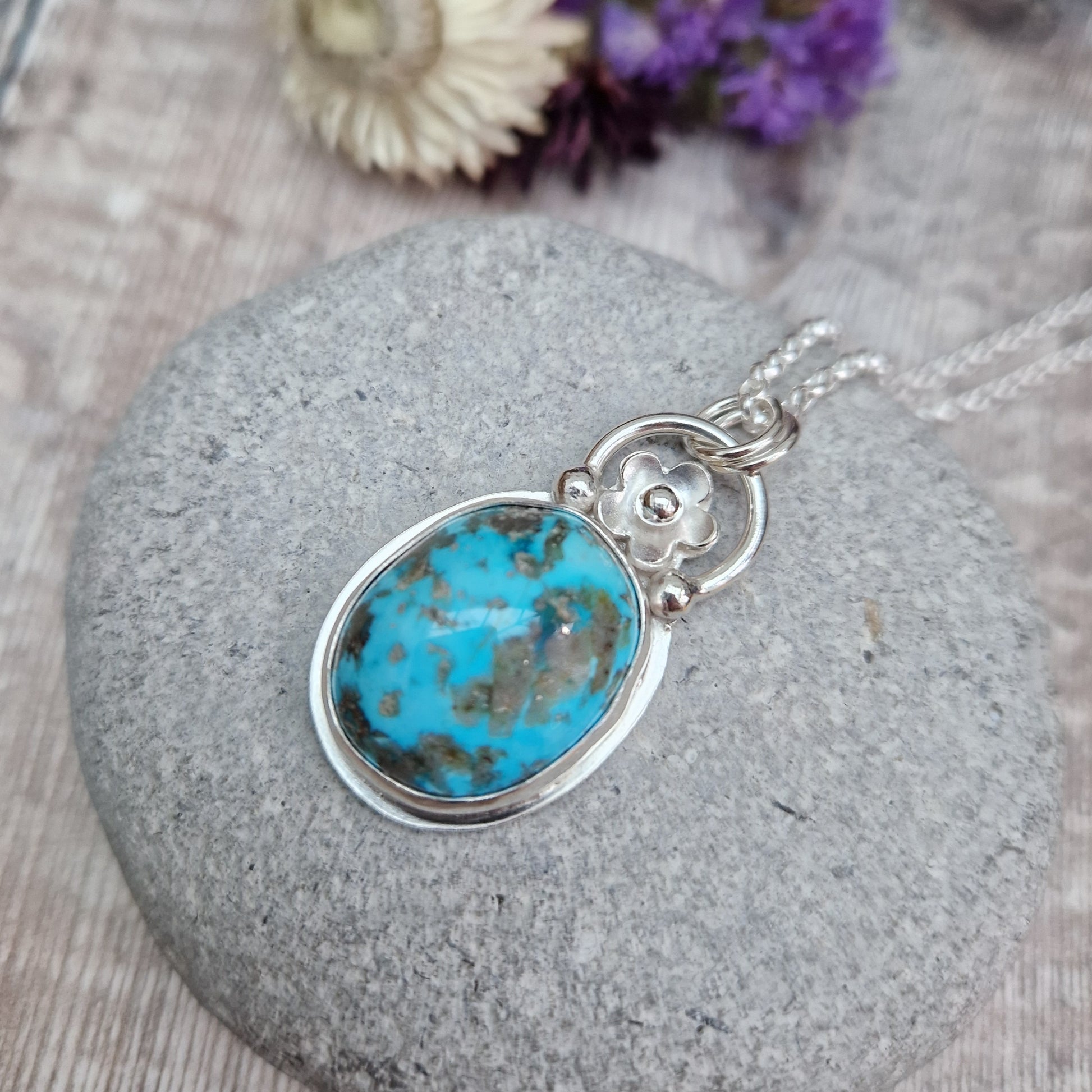 Turquoise oval gemstone pendant set in simple Sterling Silver bezel surround. Fused to the top of the bezel setting is a silver half circle with a five petaled flower in the middle, attached to silver chain via three small silver circles. Gemstone is predominantly turquoise blue with dark, almost black plume formations.