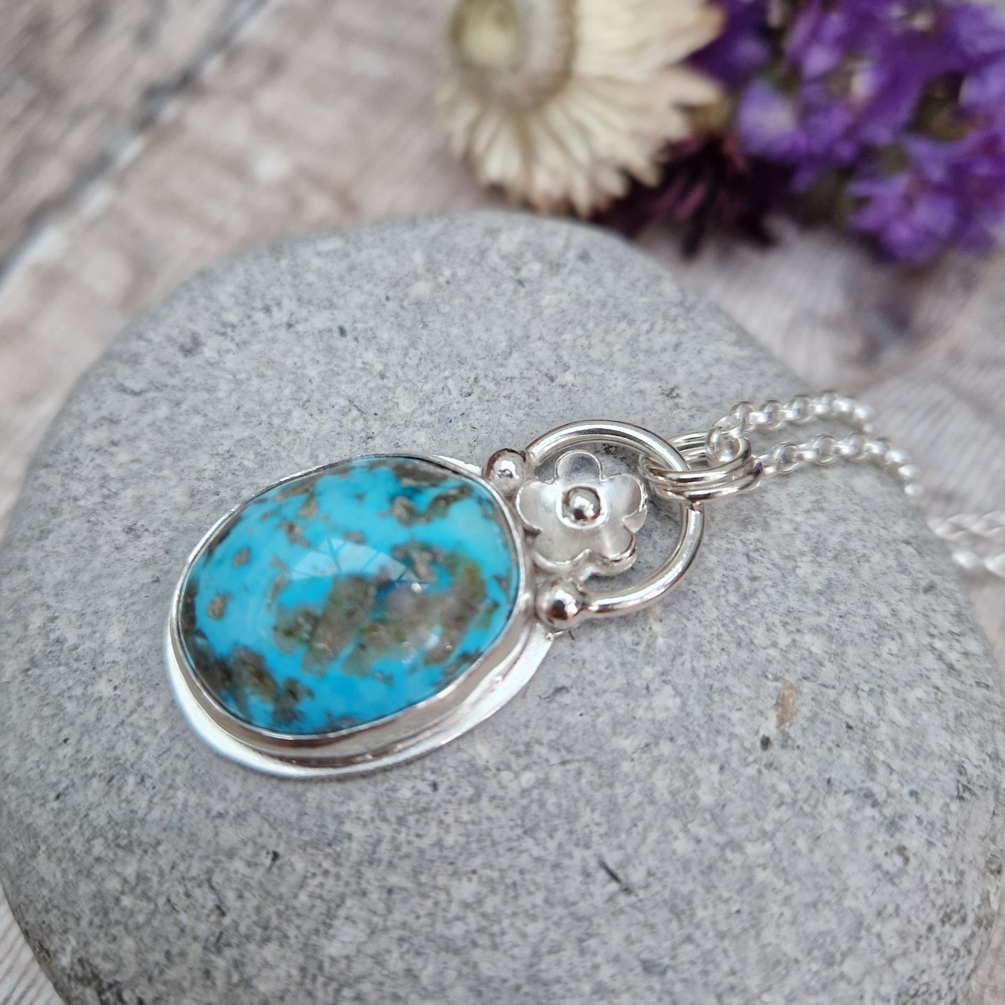 Turquoise oval gemstone pendant set in simple Sterling Silver bezel surround. Fused to the top of the bezel setting is a silver half circle with a five petaled flower in the middle, attached to silver chain via three small silver circles. Gemstone is predominantly turquoise blue with dark, almost black plume formations.