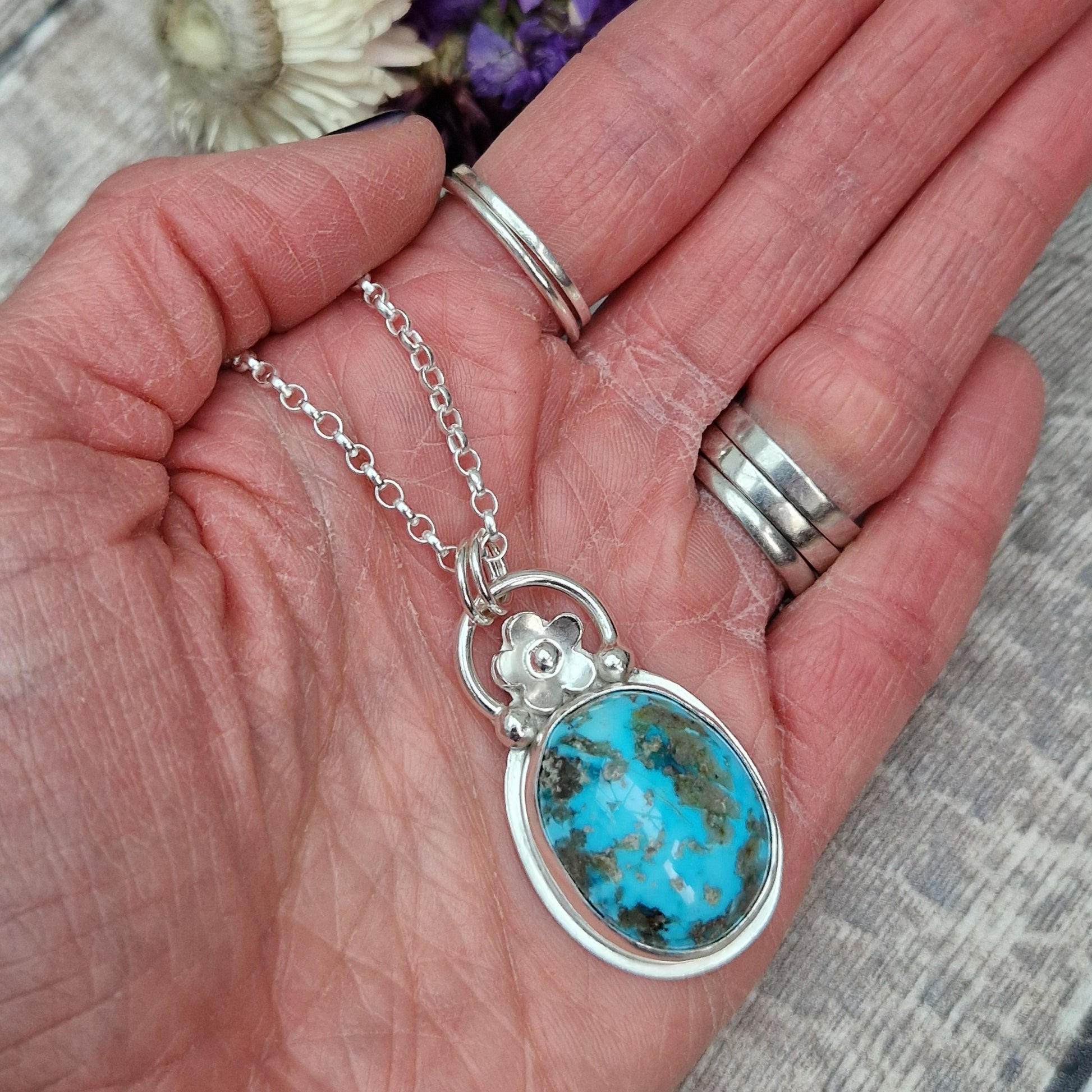 Turquoise oval gemstone pendant set in simple Sterling Silver bezel surround. Fused to the top of the bezel setting is a silver half circle with a five petaled flower in the middle, attached to silver chain via three small silver circles. Gemstone is predominantly turquoise blue with dark, almost black plume formations.