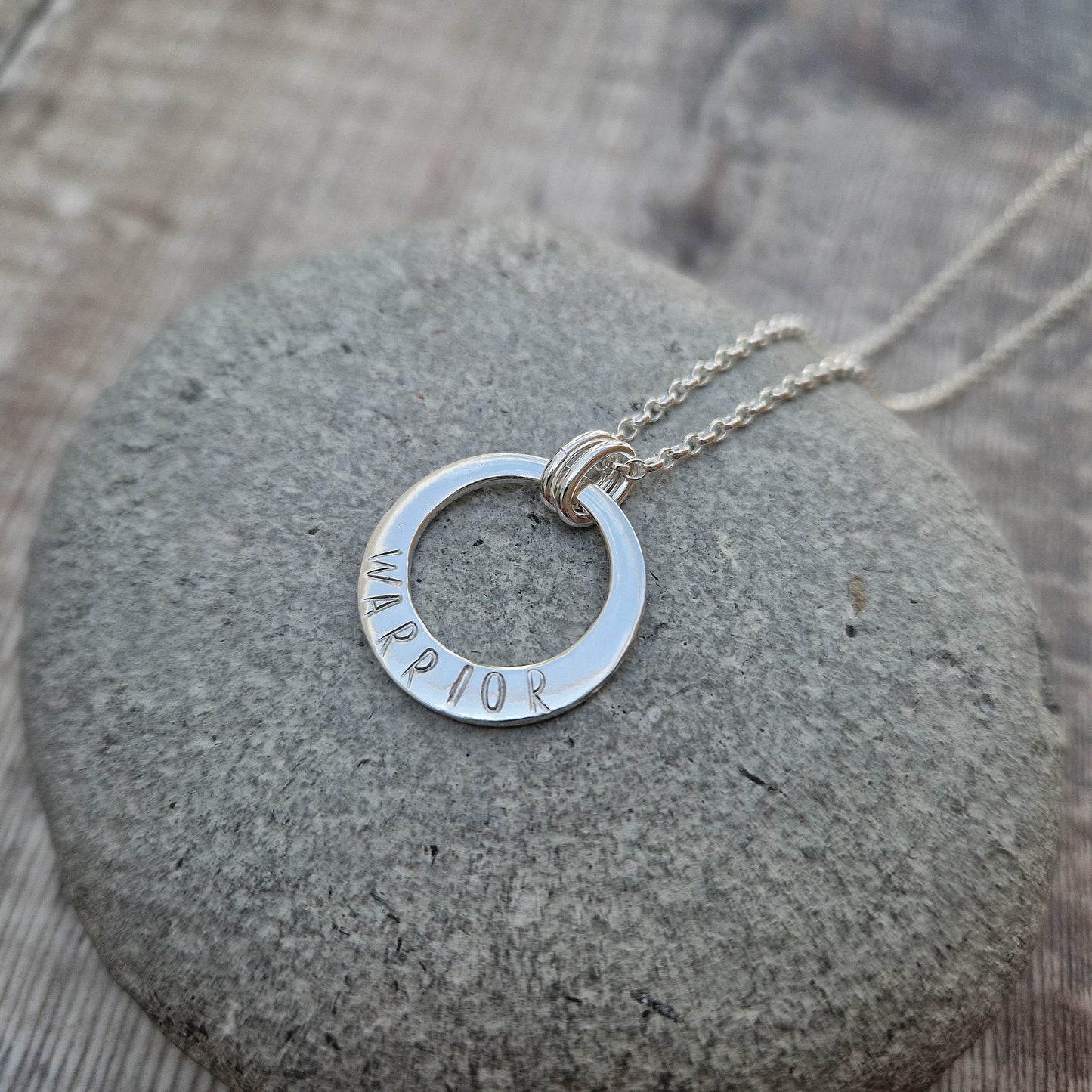 Sterling Silver ‘WARRIOR’ necklace. Silver disc with offset hole in centre, looks like a thick circle. Attached via two small silver hoops to silver chain. On thicker part of disc, inscription wording hammered into the silver reads WARRIOR in capitals. Disc measures approximately 20 mm in diameter. Chain length 16, 18 or 20 inches.