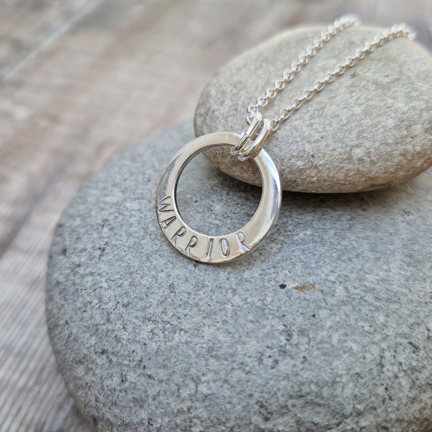 Sterling Silver ‘WARRIOR’ necklace. Silver disc with offset hole in centre, looks like a thick circle. Attached via two small silver hoops to silver chain. On thicker part of disc, inscription wording hammered into the silver reads WARRIOR in capitals. Disc measures approximately 20 mm in diameter. Chain length 16, 18 or 20 inches.