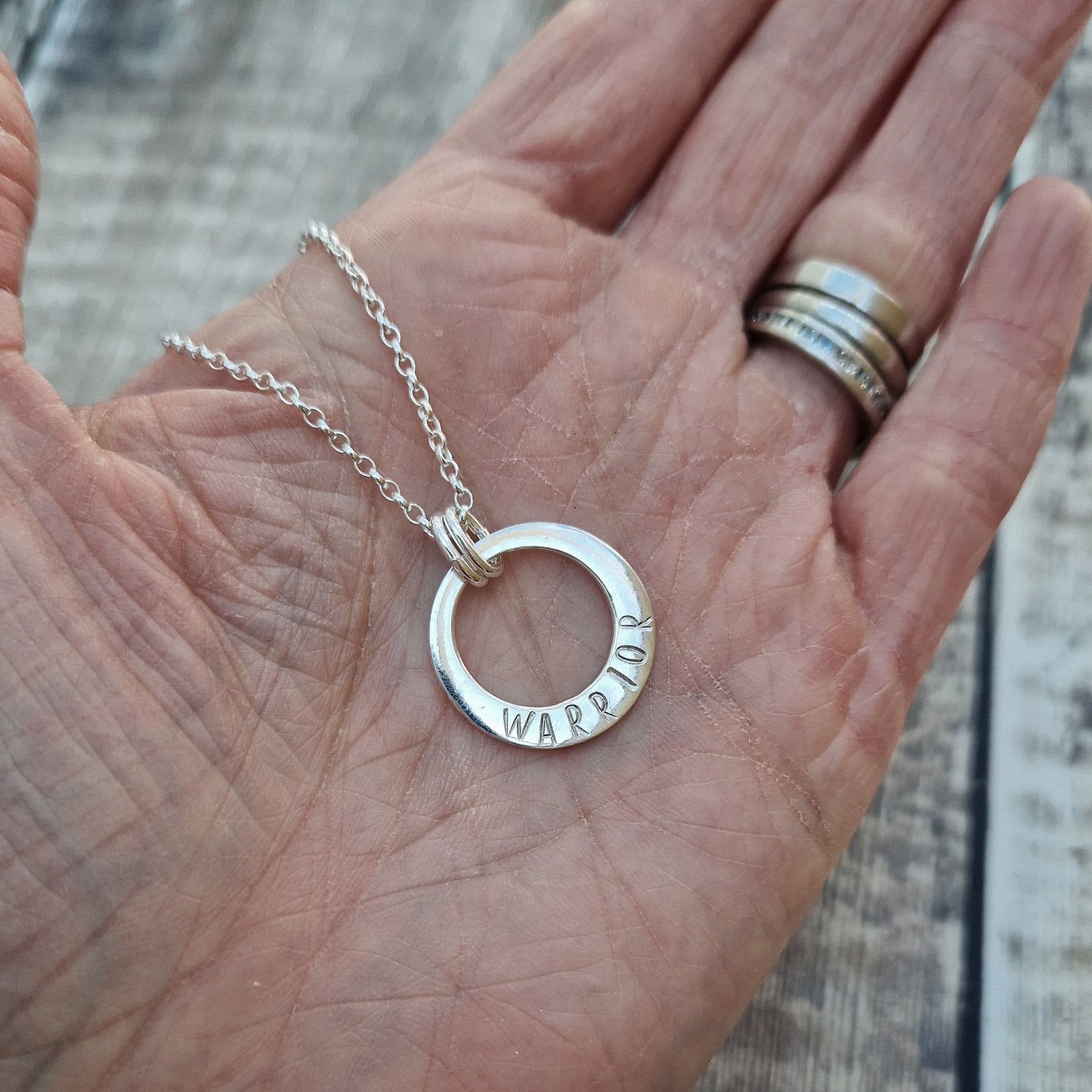 Sterling Silver ‘WARRIOR’ necklace. Silver disc with offset hole in centre, looks like a thick circle. Attached via two small silver hoops to silver chain. On thicker part of disc, inscription wording hammered into the silver reads WARRIOR in capitals. Disc measures approximately 20 mm in diameter. Chain length 16, 18 or 20 inches.