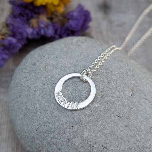 A silver circle necklace with the word "WARRIOR" hand-stamped on the lower part of the pendant, resting on a smooth grey stone. The chain is draped over the stone, with a blurred background featuring dried purple flowers.