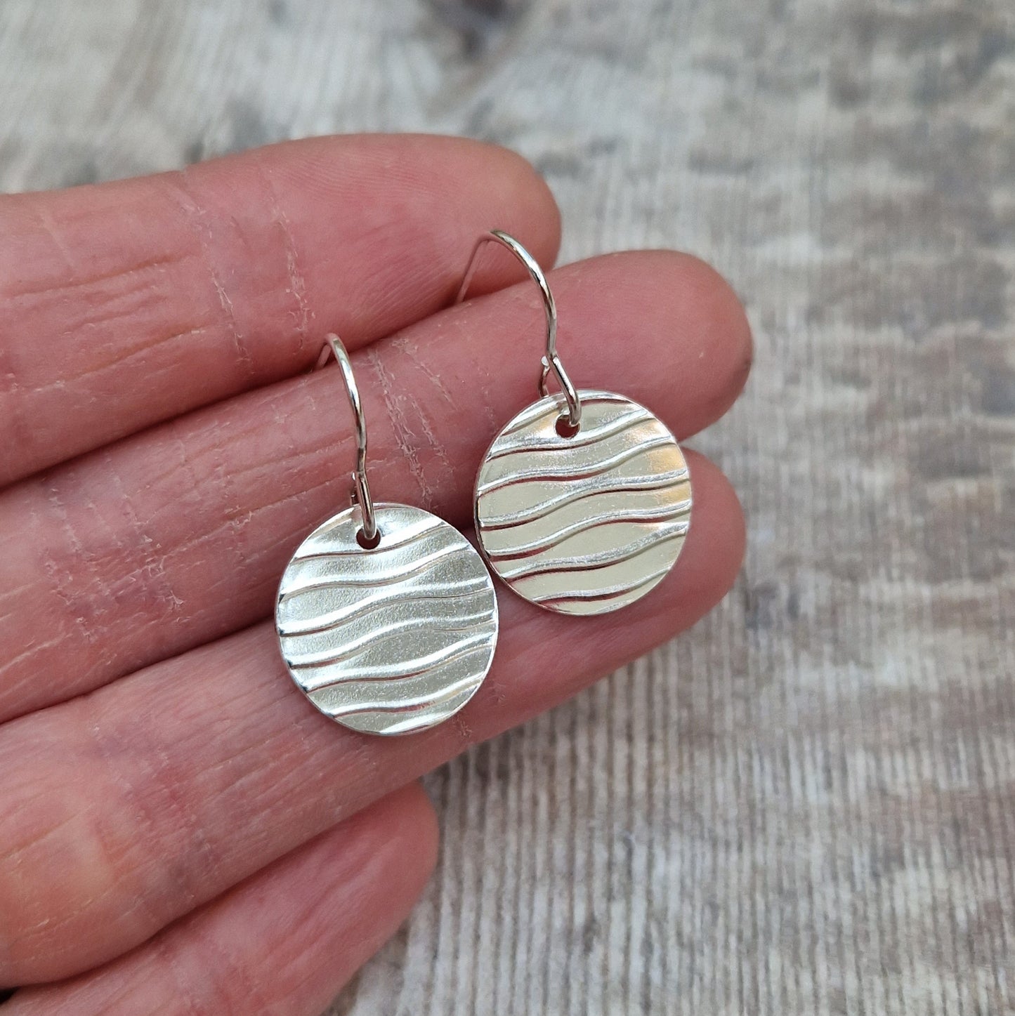 Each earring is a sterling silver disc stamped with a wavy line pattern. The wavy lines are spaced apart and raised from the disc, suspended from silver earring wire.