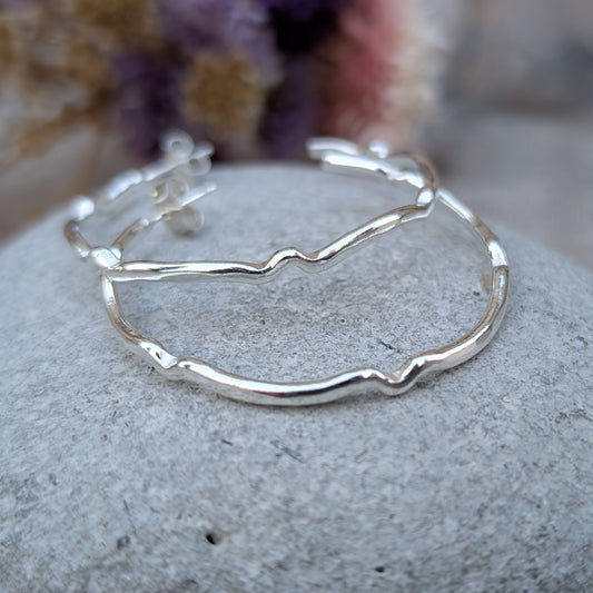 Each earring is a silver hoop attached to stud bar. The hoop is irregular shaped in repeating waves, represented as small bumps at intervals. Polished silver finish.