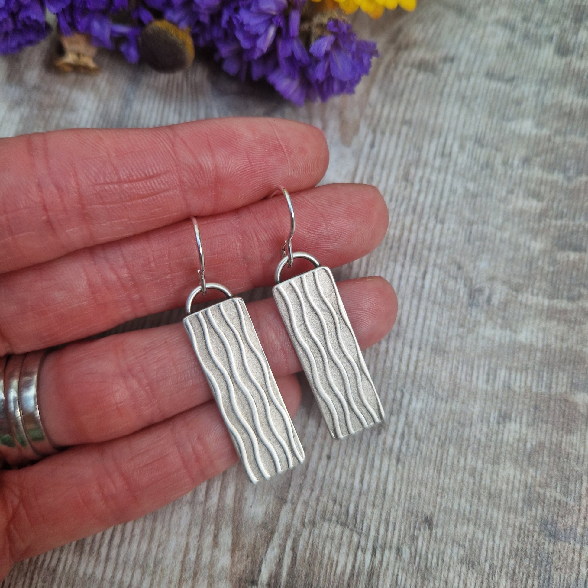 Each earring is an Oxidised Sterling Silver rectangular shaped pendant suspended from silver earring wire. Stamped on the rectangular shape are vertical wavy lines going the length of the pendant. Parts of the pattern are darker and lighter.