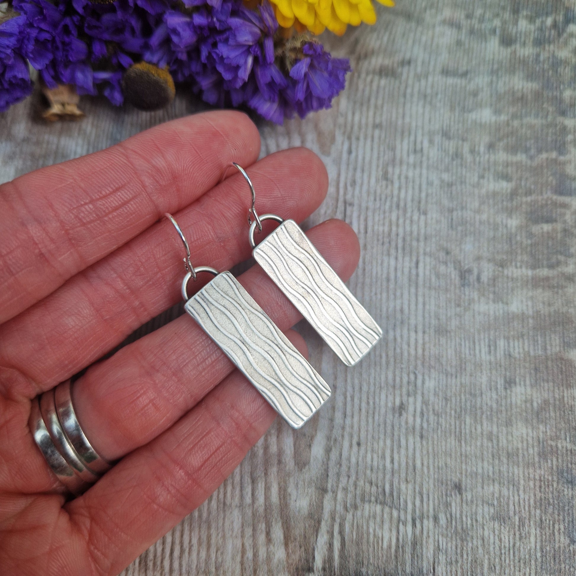 Each earring is an Oxidised Sterling Silver rectangular shaped pendant suspended from silver earring wire. Stamped on the rectangular shape are vertical wavy lines going the length of the pendant. Parts of the pattern are darker and lighter.