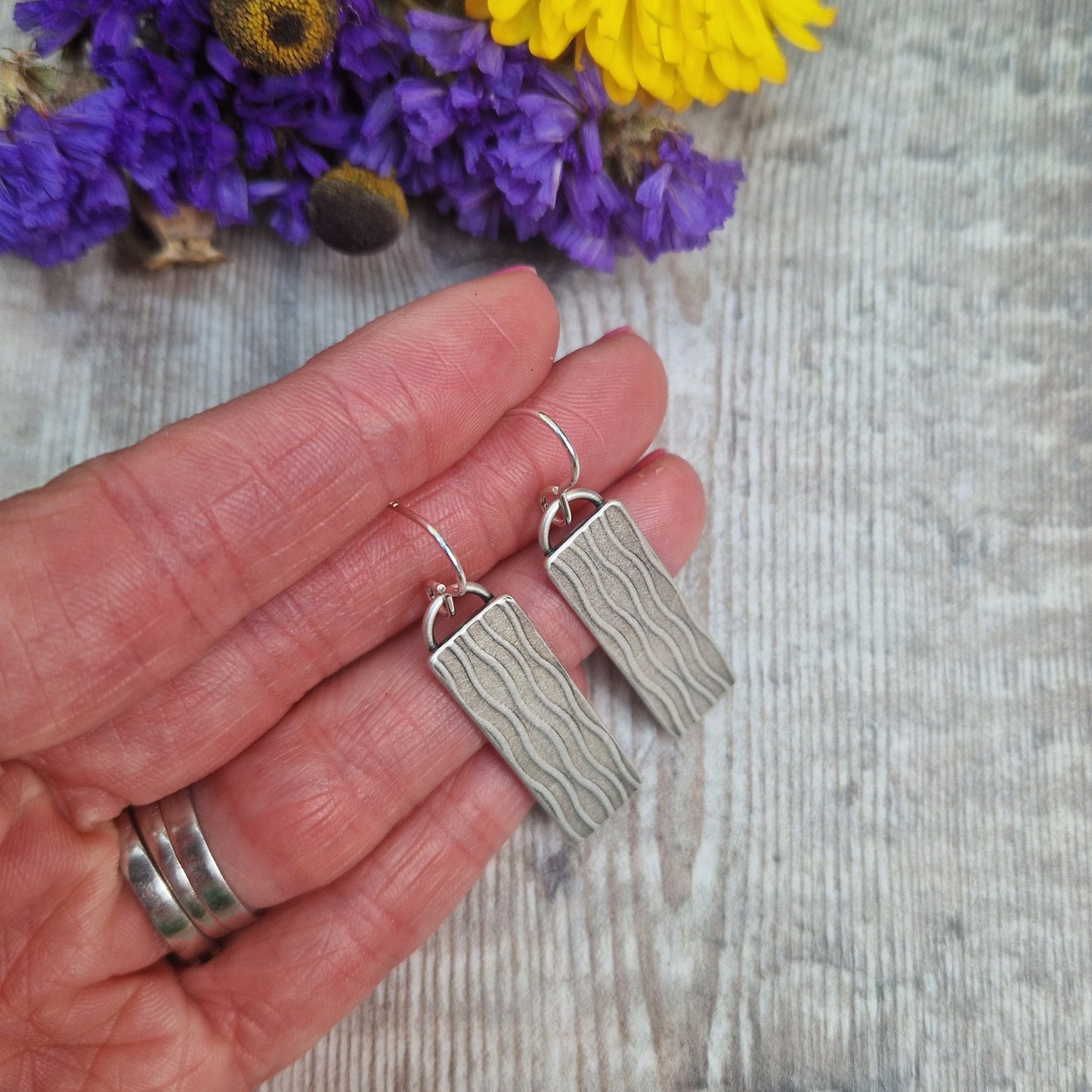 Each earring is an Oxidised Sterling Silver rectangular shaped pendant suspended from silver earring wire. Stamped on the rectangular shape are vertical wavy lines going the length of the pendant. Parts of the pattern are darker and lighter.