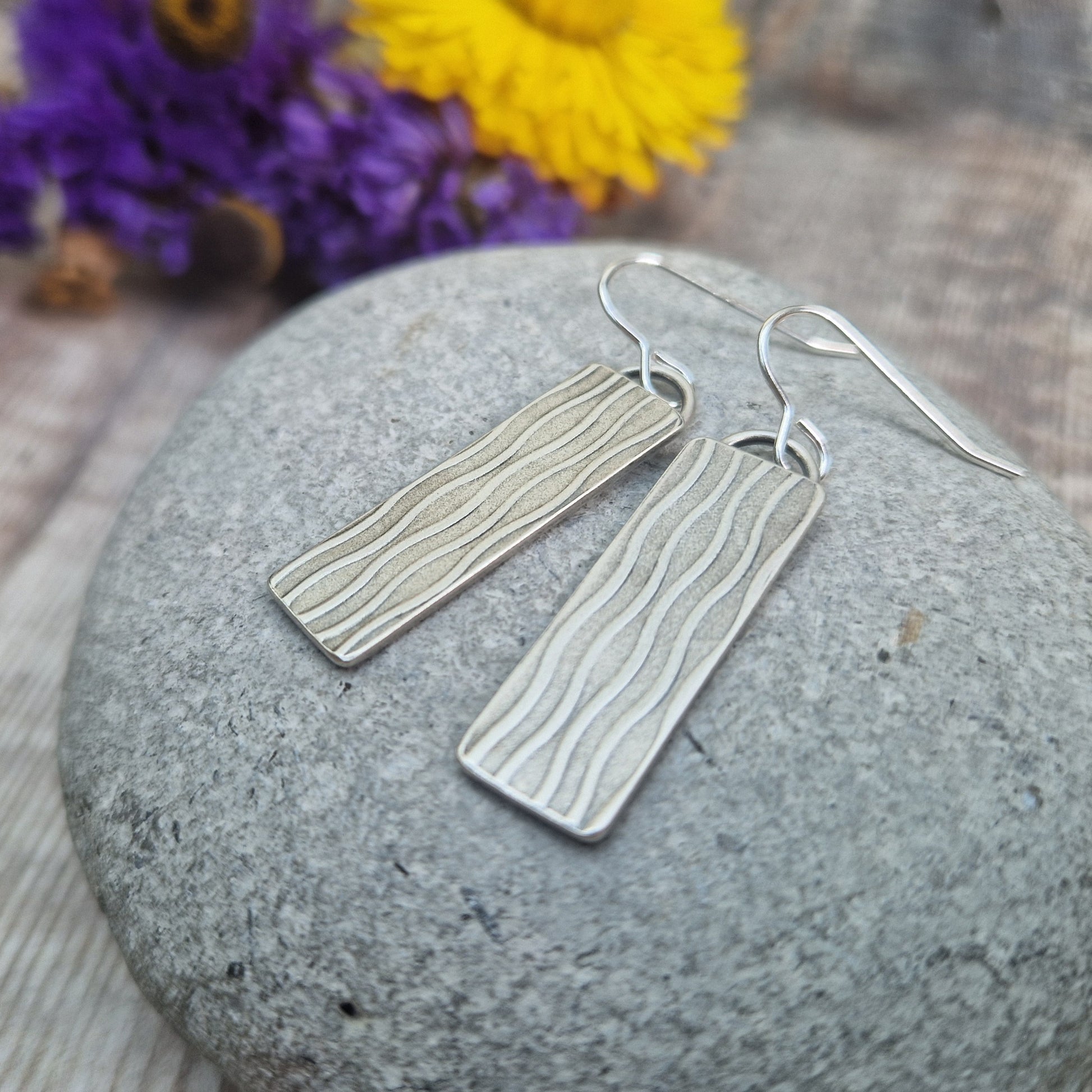 Each earring is an Oxidised Sterling Silver rectangular shaped pendant suspended from silver earring wire. Stamped on the rectangular shape are vertical wavy lines going the length of the pendant. Parts of the pattern are darker and lighter.